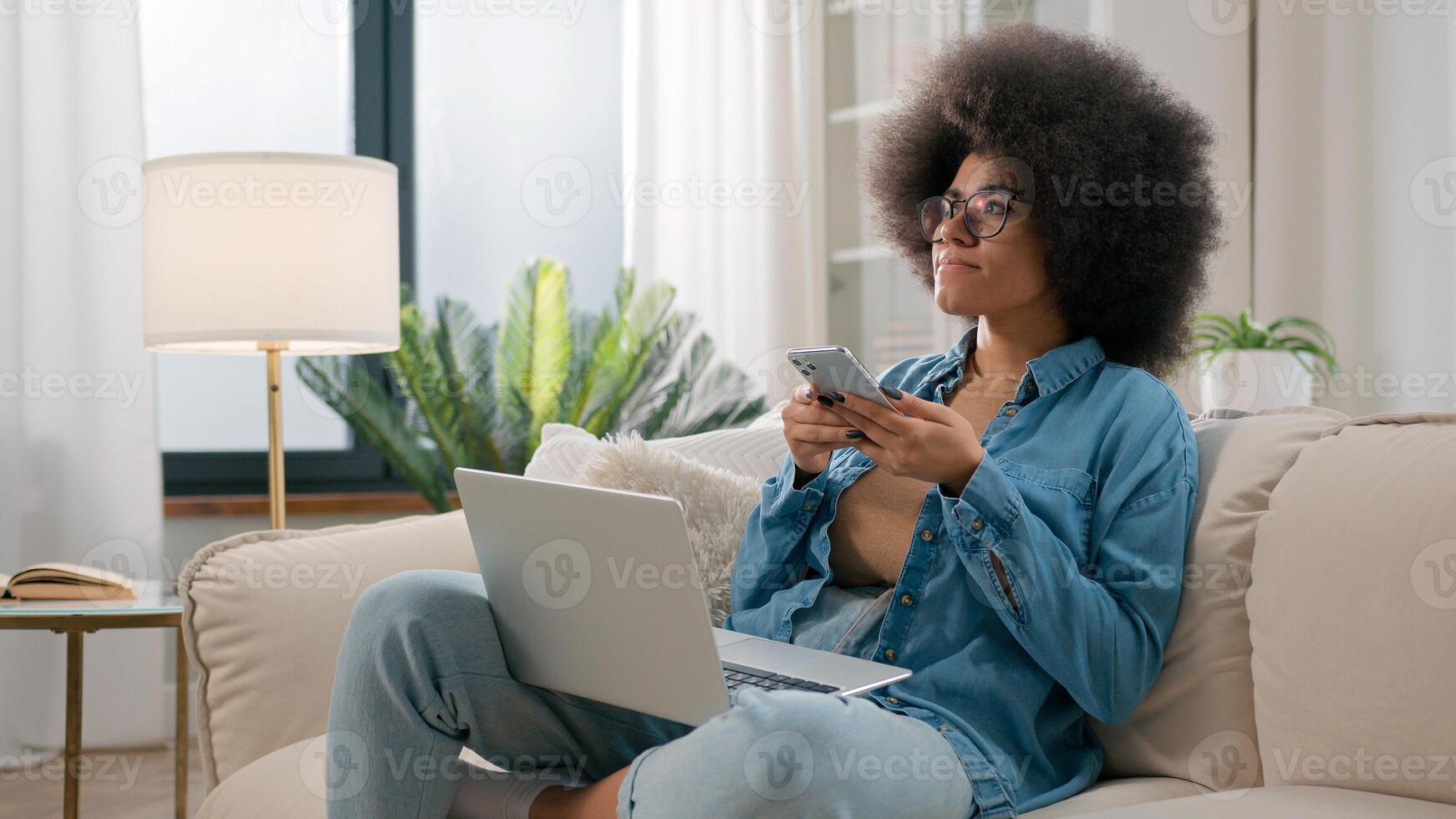 africano americano mujer confuso perplejo pensativo étnico niña con ordenador portátil en sofá hojeada móvil teléfono pensando problema solución escoger comida entrega descanso en distante trabajo desplazamiento teléfono inteligente a hogar foto