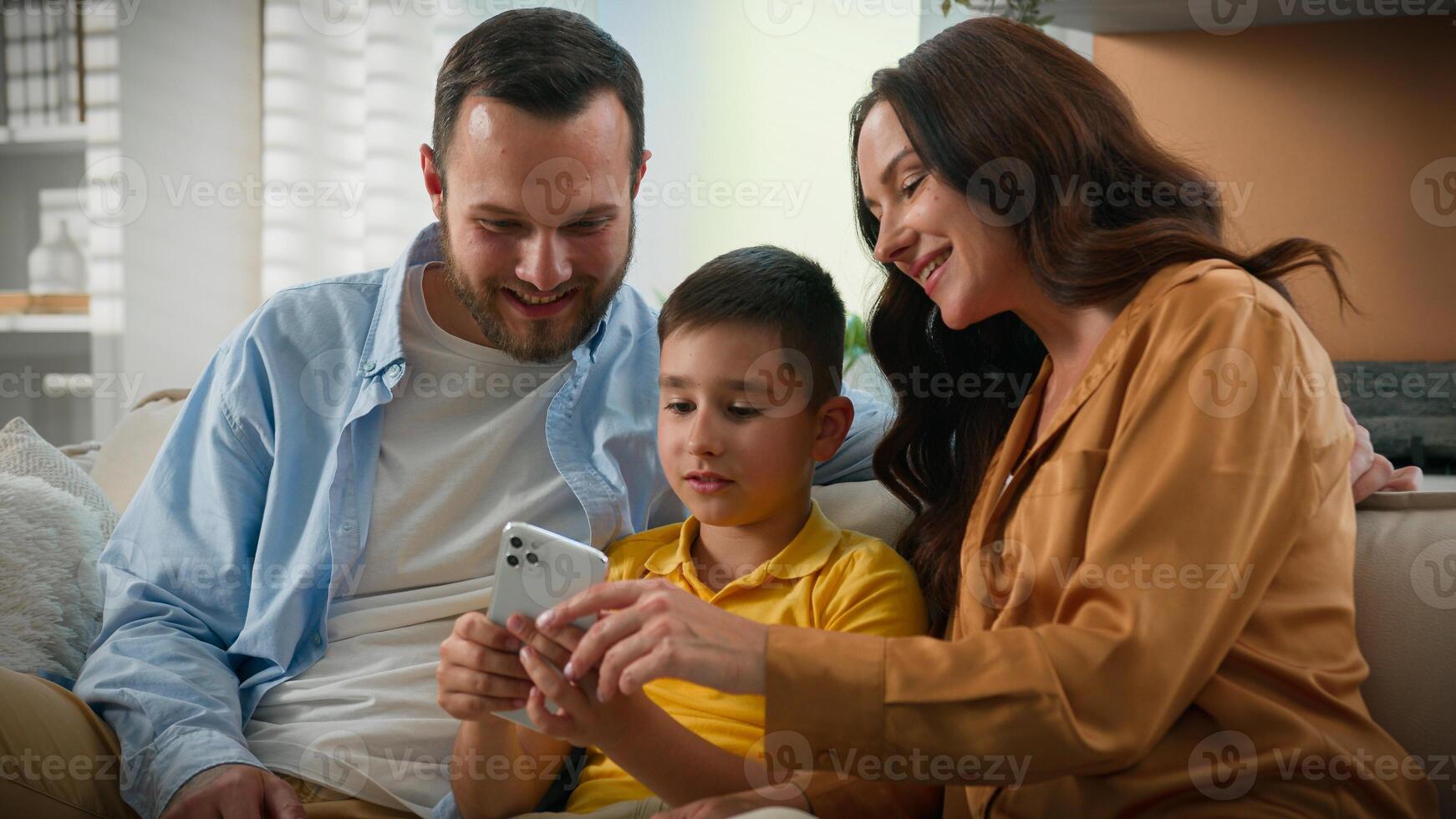 Happy Caucasian family man woman father mother kid boy child son making purchase in online toy store browsing social network make order in Internet on phone smartphone on couch home parenthood concept photo