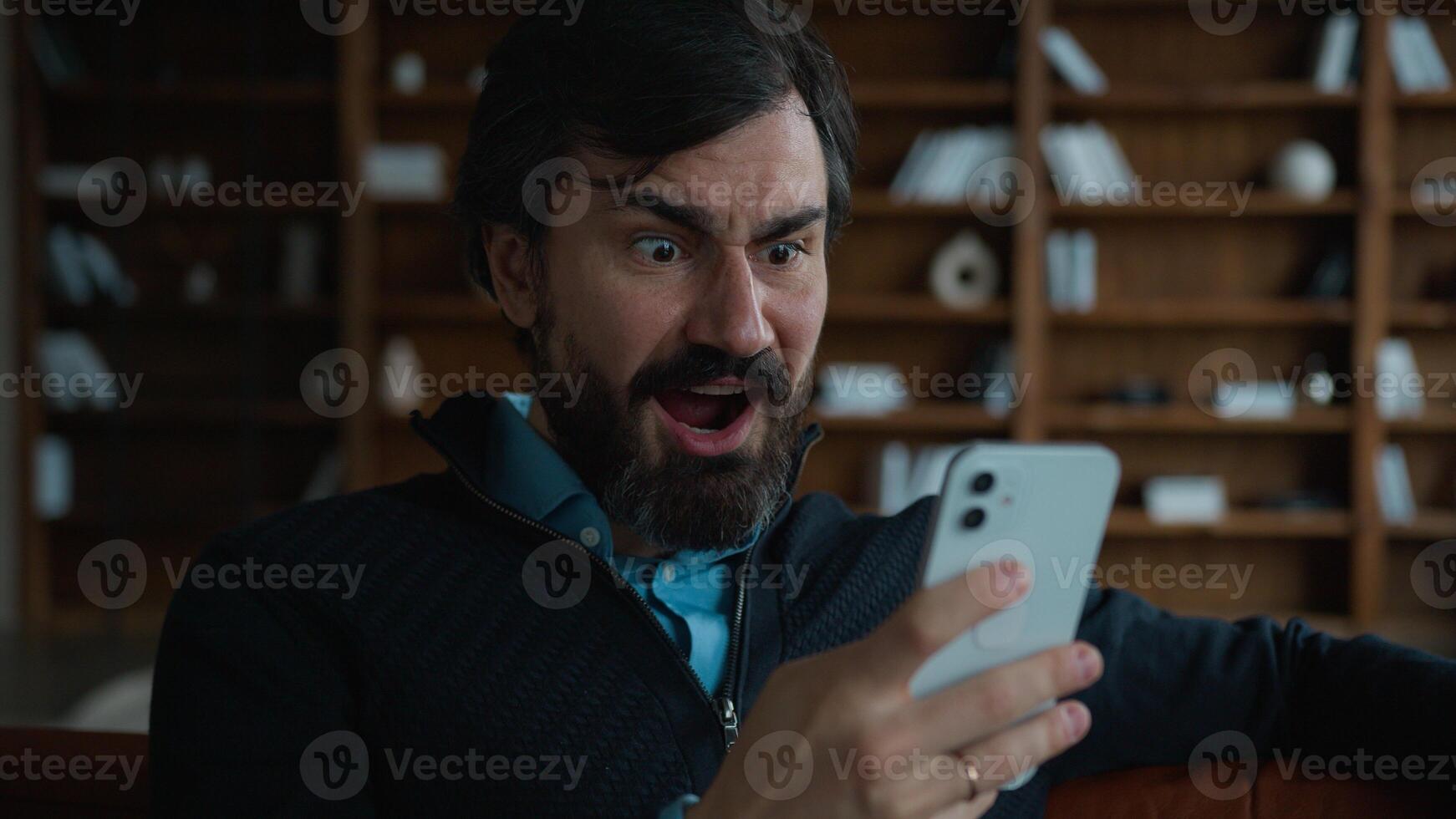 Shocked amazed excited 50s mature middle aged Caucasian business man male businessman hold smartphone use mobile cell phone receive amazing news win game victory online bet offer prize winning indoors photo