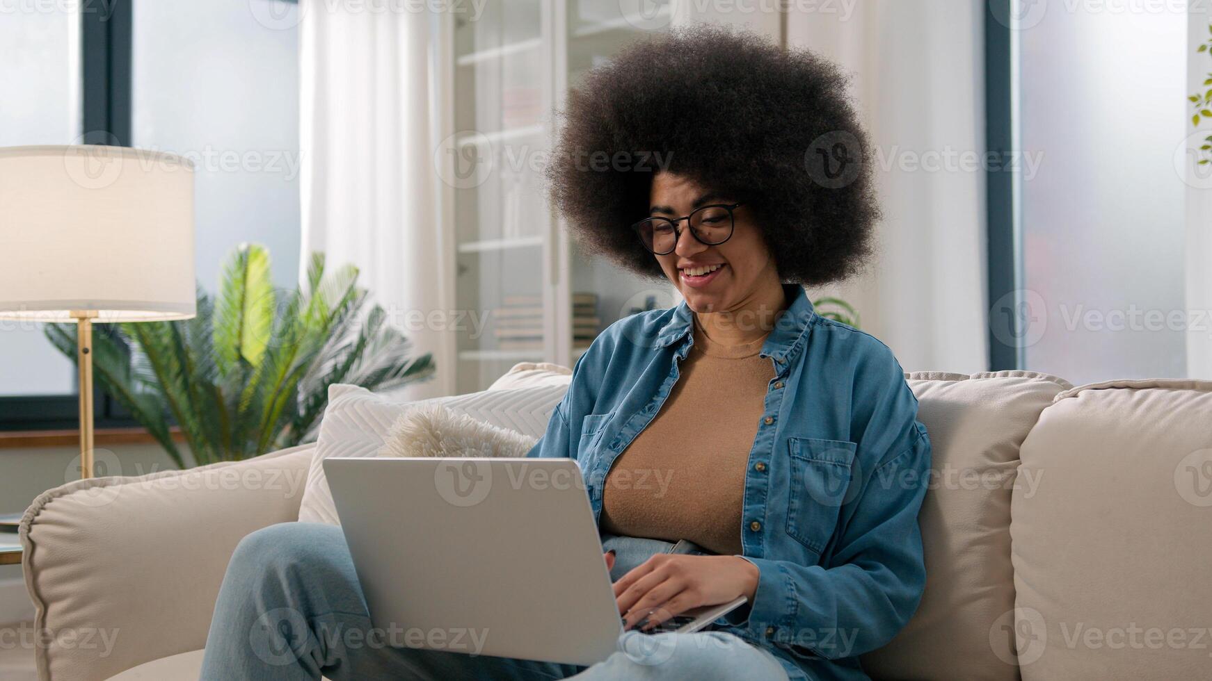 Young African American girl working distant from home with laptop at sofa ethnic woman freelancer typing email on computer online job using web service for e-learning study on couch user browsing net photo