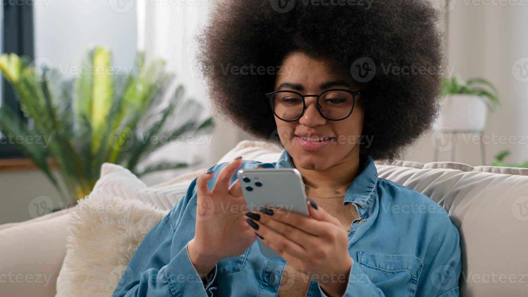 Relaxed calm African American ethnic woman typing swipe touchscreen scrolling mobile phone check social media cellphone app girl choosing purchase shopping in online e-commerce application at home photo