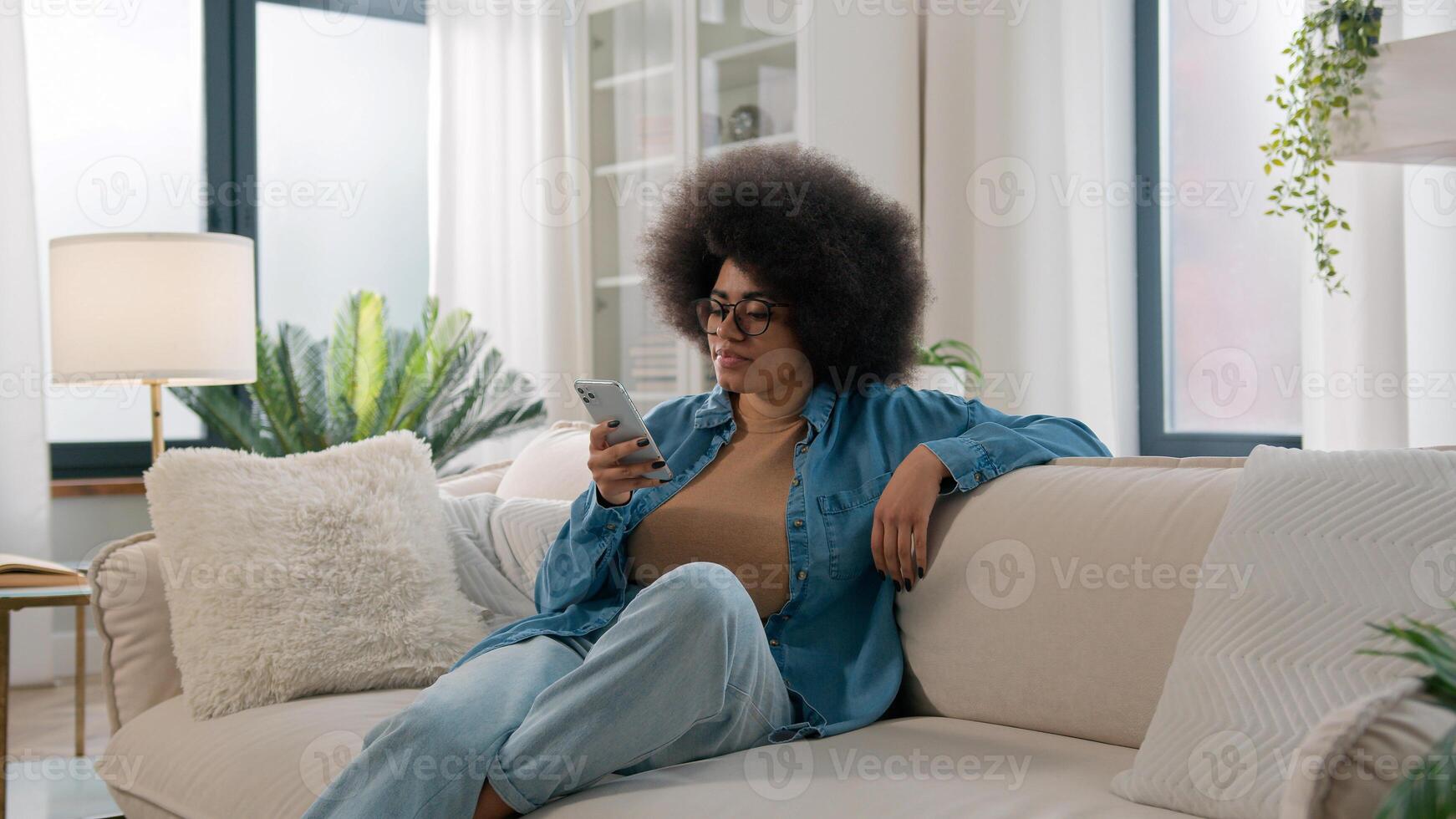Relaxed calm African American ethnic woman typing swipe touchscreen scrolling mobile phone check social media cellphone app girl choosing purchase shopping in online e-commerce application at home photo