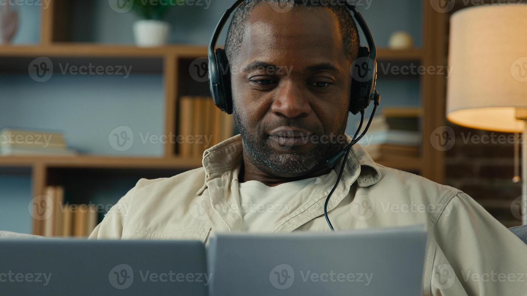 African American man male businessman sales manager freelancer homeowner working on laptop at home using headset checking documents paying bills browsing online information paperwork entering data photo