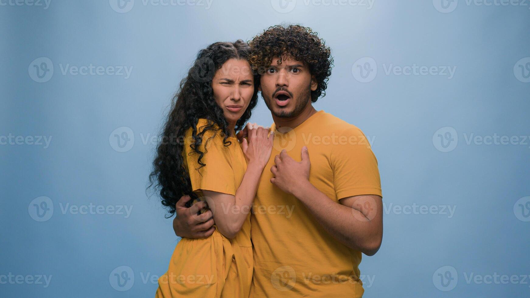 Multiracial shocked scared afraid couple amazed shocked Hispanic woman hiding behind Indian man astonishment wonder scare terrible horror ugly reaction blue studio background shock astonish fright photo