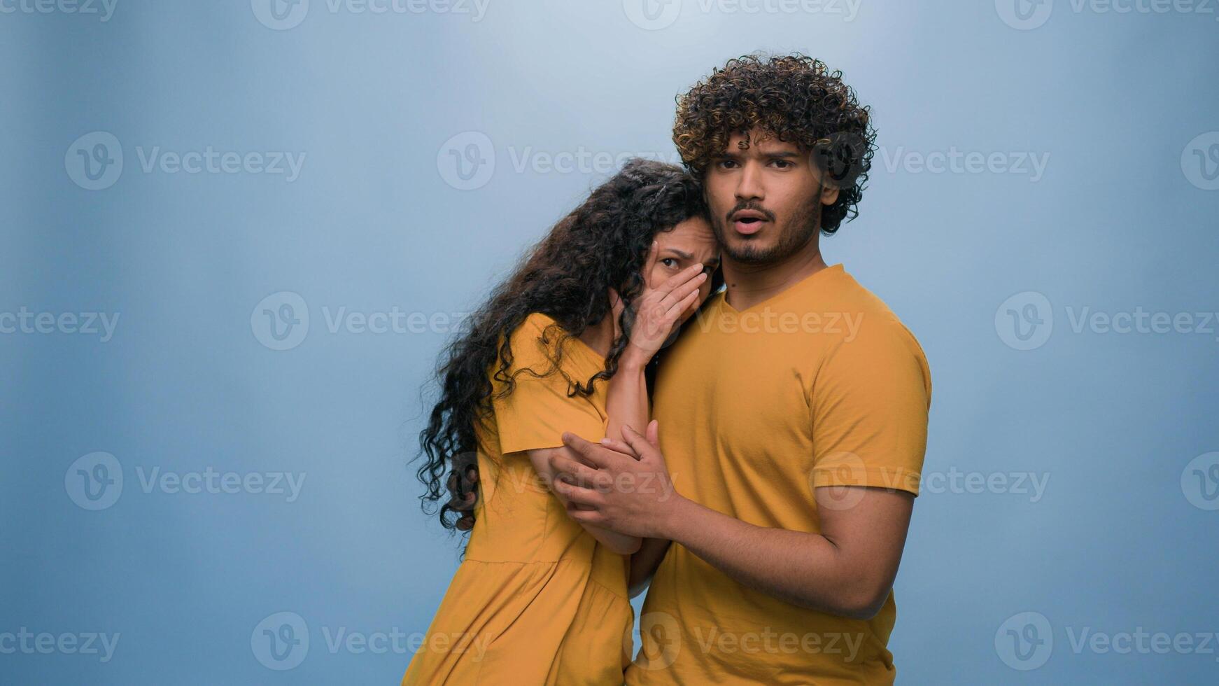 Multiracial shocked scared afraid couple amazed shocked Hispanic woman hiding behind Indian man astonishment wonder scare terrible horror ugly reaction blue studio background shock astonish fright photo