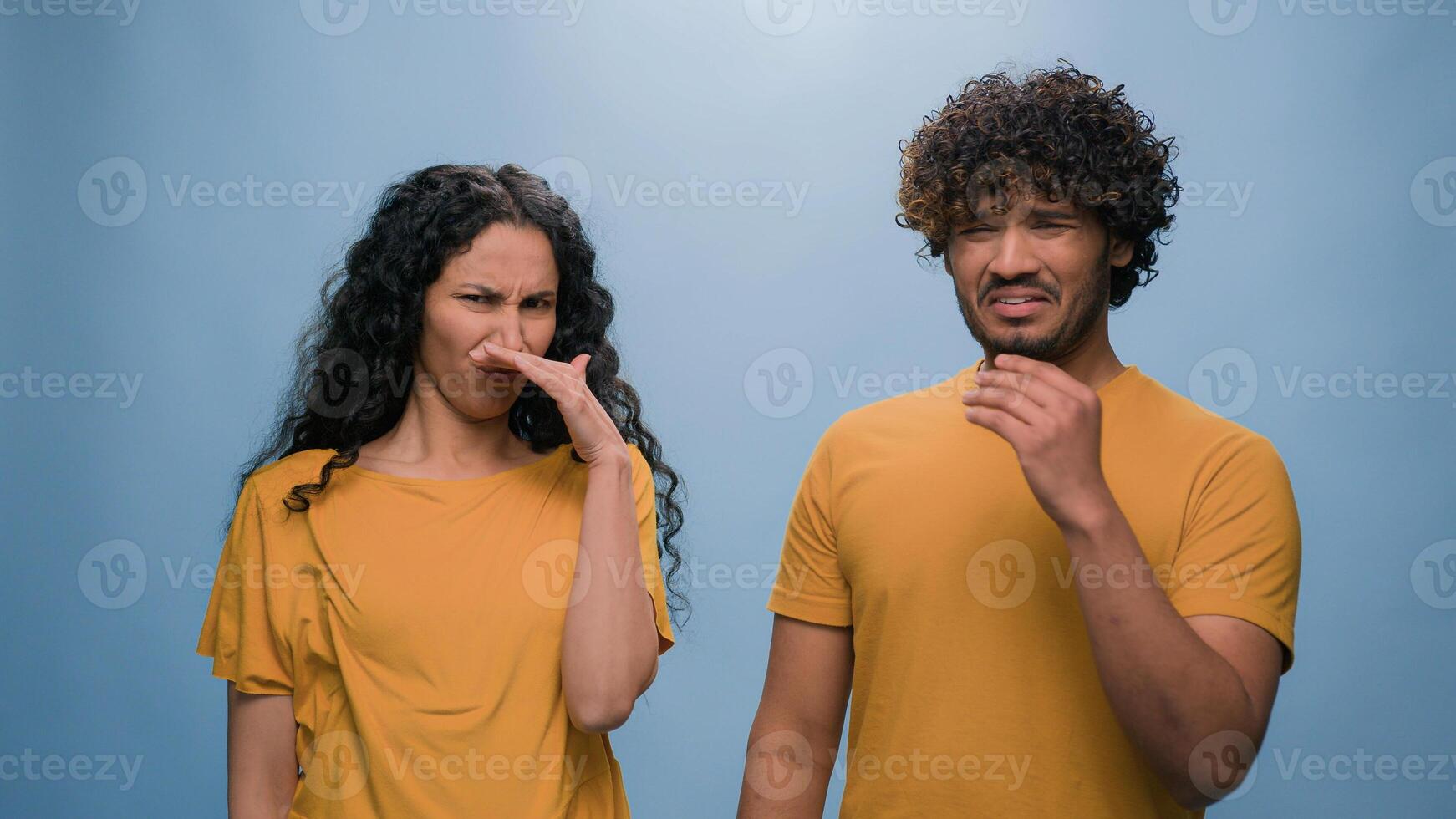 Bad smell reaction multiracial diverse couple Indian Arabian man and Hispanic woman cover noses suffer disgust stink stinky smelly aroma frowning sniffing unpleasant odor at blue studio background photo