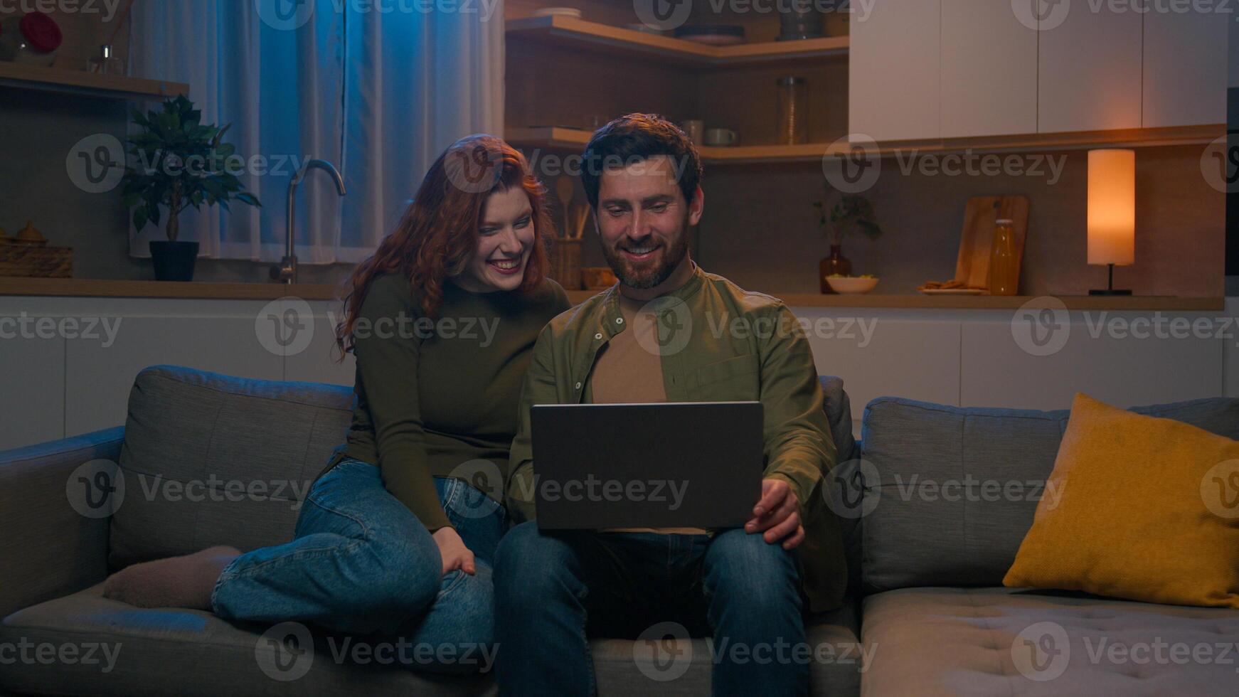 Happy smiling Caucasian man husband woman wife married couple surfing computer on couch home relaxing together romantic evening browsing media network using laptop order delivery buy in online store photo