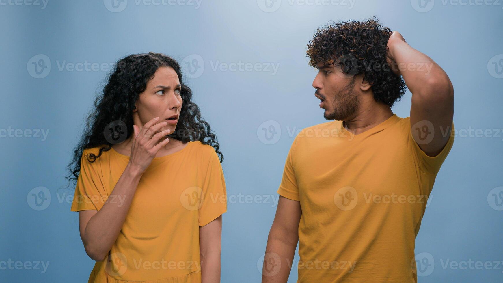 Multiracial shocked scared afraid couple amazed shocked Hispanic woman hiding behind Indian man astonishment wonder scare terrible horror ugly reaction blue studio background shock astonish fright photo