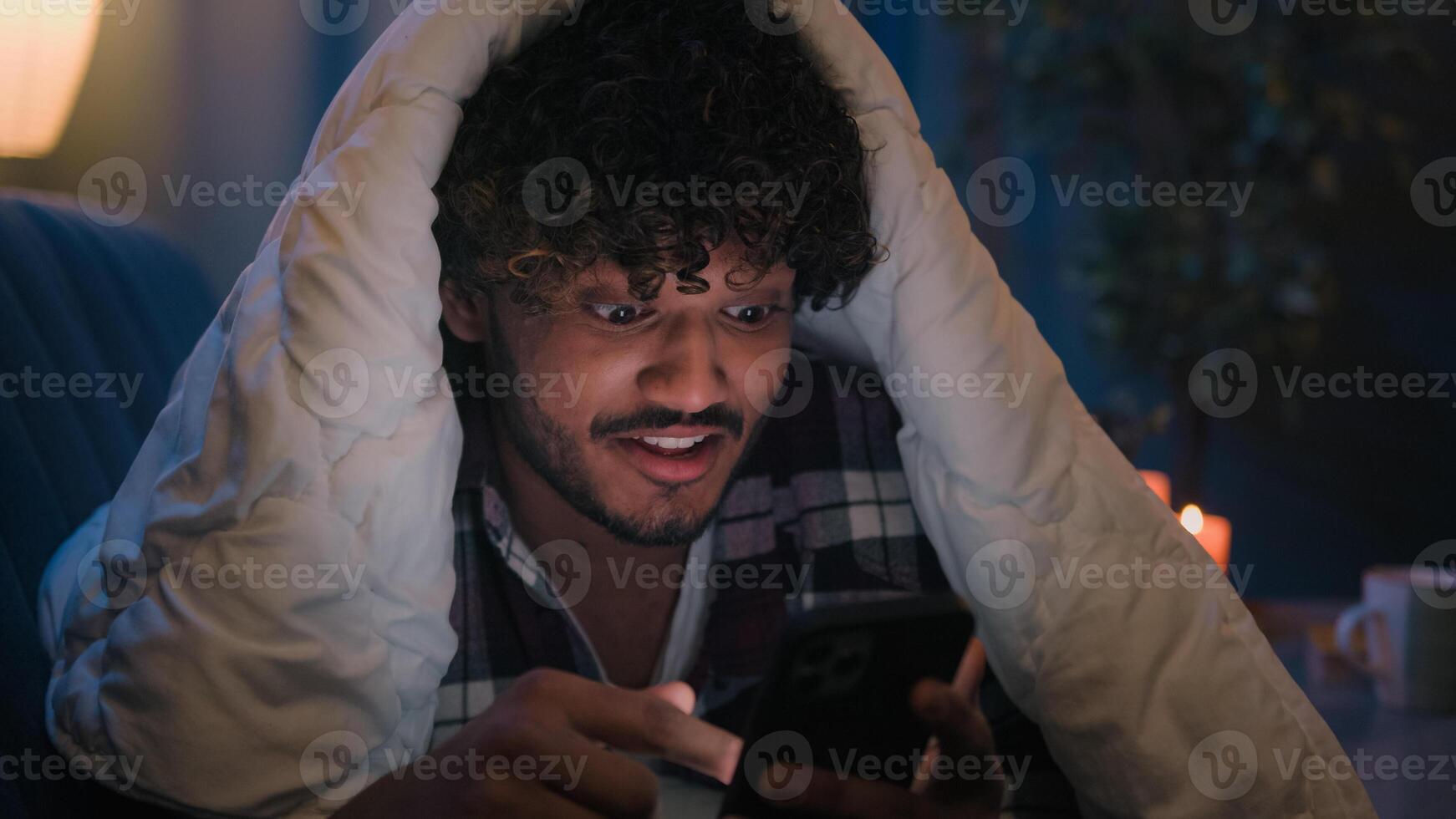 Smiling guy Indian man Arabian male at night evening home on couch under blanket cover with duvet looking mobile phone wow surprised amazed reading news gadget addict smartphone social media scrolling photo