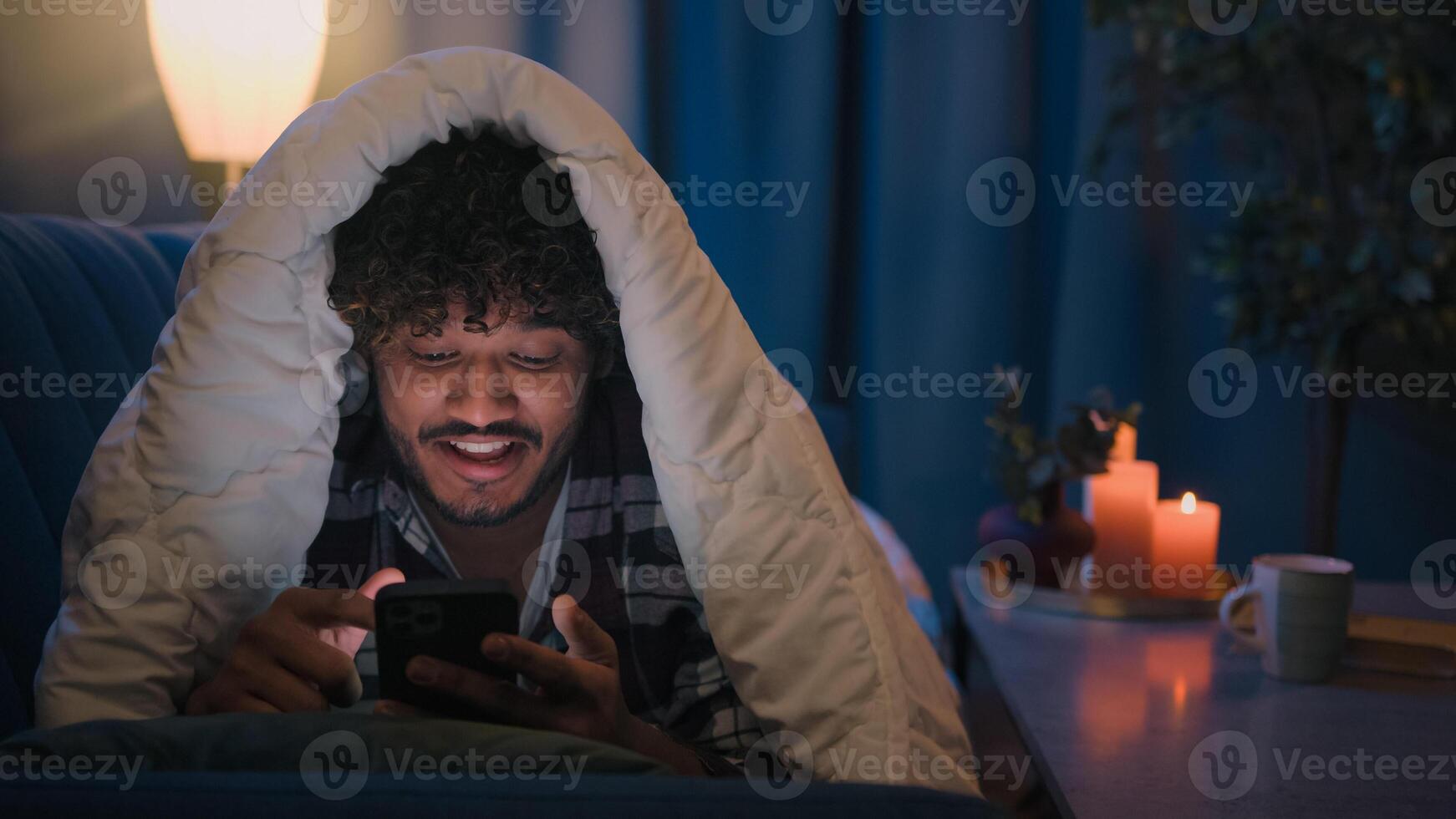 Smiling guy Indian man Arabian male at night evening home on couch under blanket cover with duvet looking mobile phone wow surprised amazed reading news gadget addict smartphone social media scrolling photo
