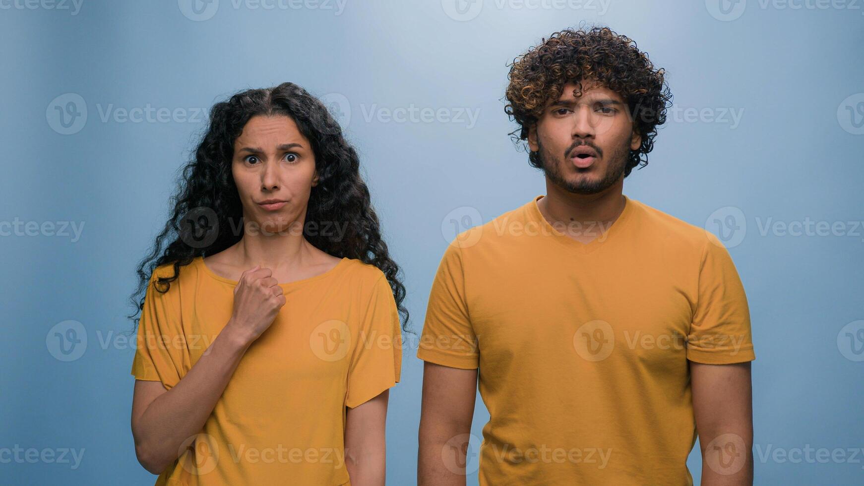 Multiracial shocked scared afraid couple amazed shocked Hispanic woman hiding behind Indian man astonishment wonder scare terrible horror ugly reaction blue studio background shock astonish fright photo