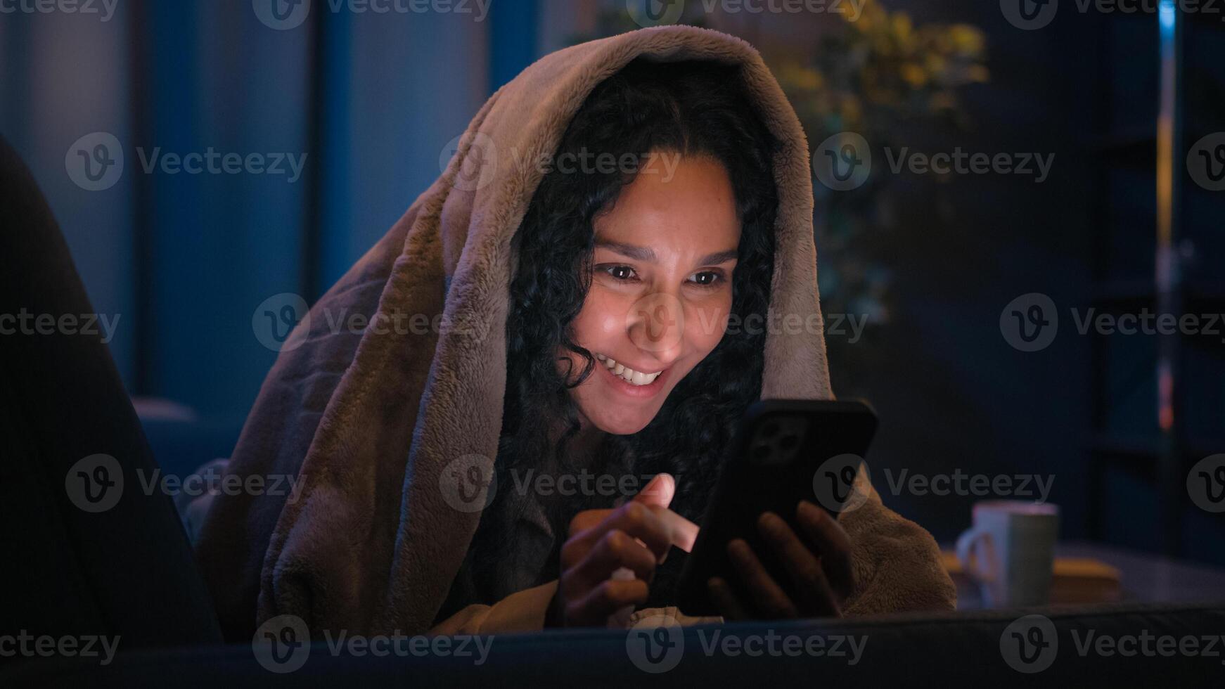 Hispanic woman girl using smartphone under blanket on bed couch reading social media funny network laugh fun laughing enjoy humorous mobile phone illuminated by bright monitor at night gadget addict photo