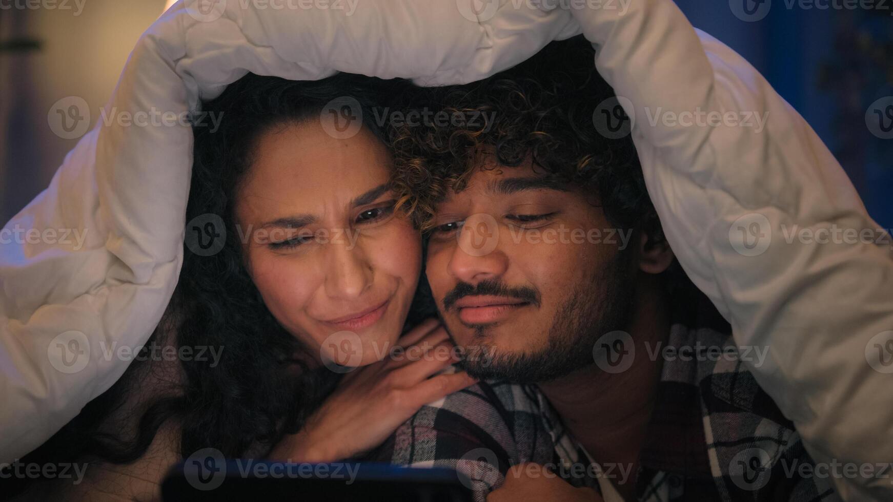 Multiracial couple man and woman happy cover with blanket under duvet at night evening home using mobile phone online browsing talking discuss call shopping with smartphone family gadget addict photo