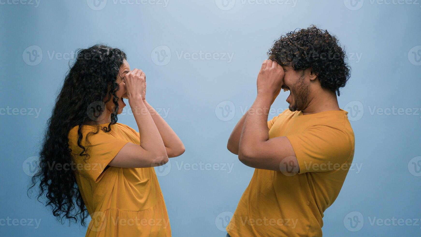 Portrait in blue background studio funny couple Indian man Arabian woman Hispanic friends girl guy having fun looking through hands make circles shape binoculars glasses goggles eyeglasses spy vision photo