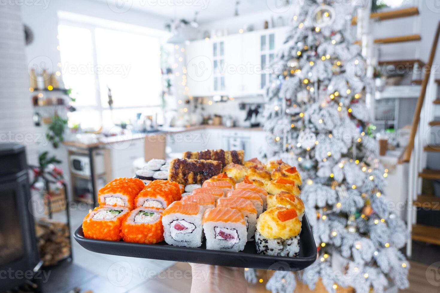 Delivery of sushi and rolls set is hold in hand in the festive interior of house with a white Christmas tree for Christmas and New year. Food delivery for the holidays photo