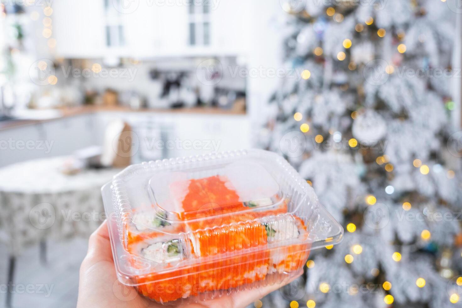 Delivery of sushi and rolls set is hold in hand in the festive interior of house with a white Christmas tree for Christmas and New year. Food delivery for the holidays photo