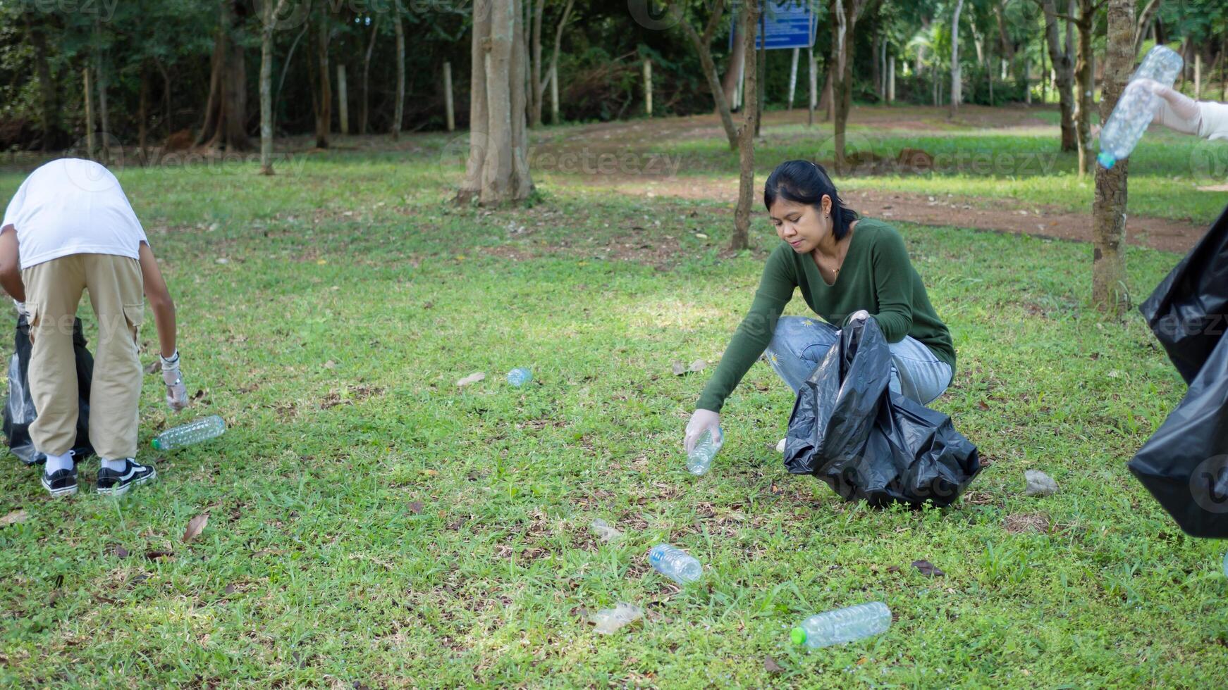 A group of dedicated young volunteers are taking part in an environmental cleaning activity in a park. Volunteers are holding big black bags. People are picking up trash and plastic bottles in a park. photo