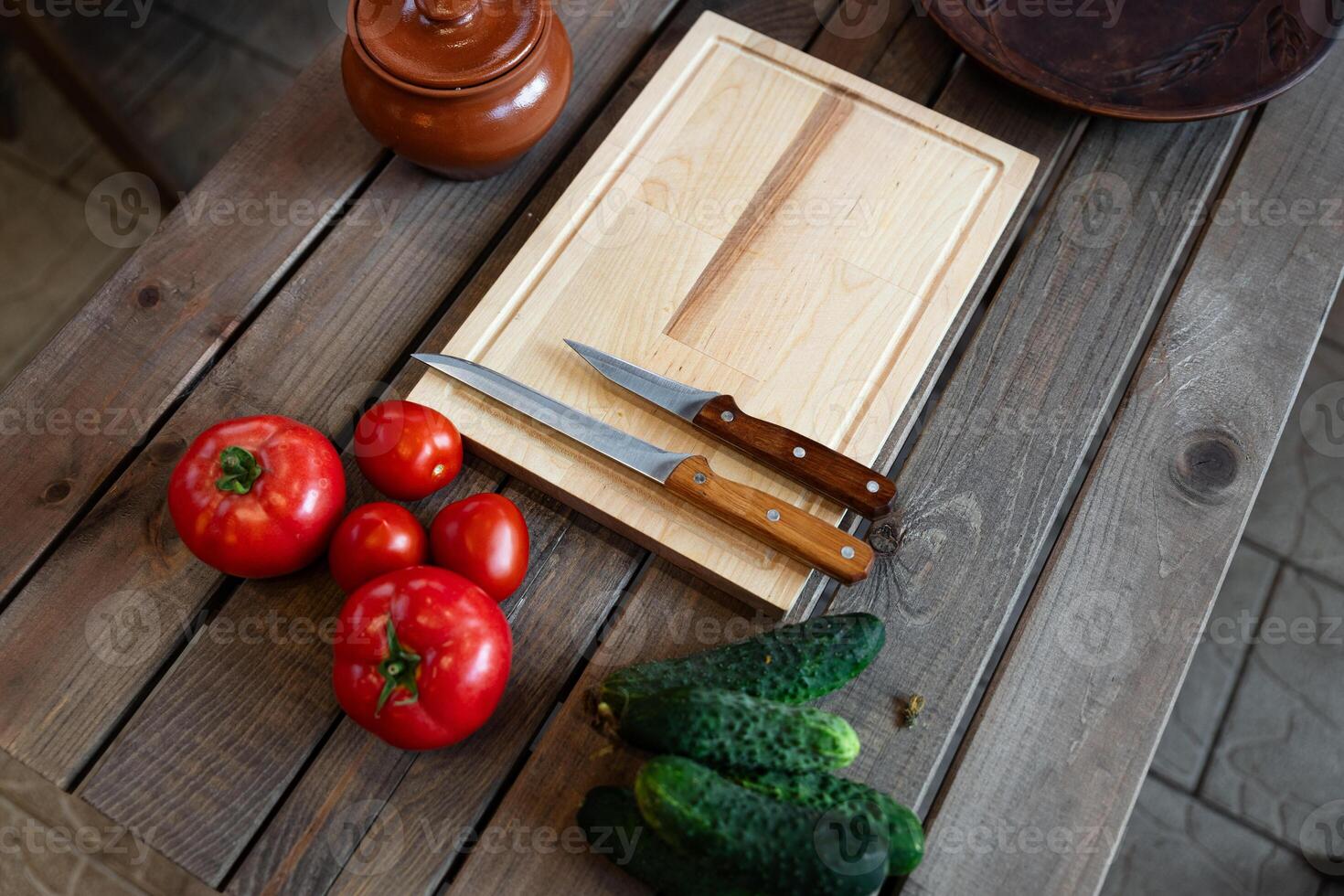 Wooden table with some fresh vegetables and kitchen items. There are several tomatoes and cucumbers placed next to a wooden cutting board. photo