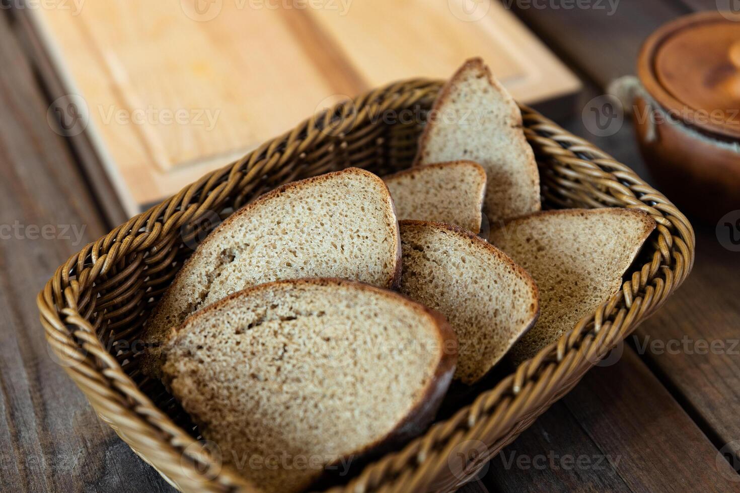 Basket filled with slices of bread. The basket is woven and sits on a wooden table. There are also two ceramic pots with lids nearby, adding a rustic feel to the scene. photo
