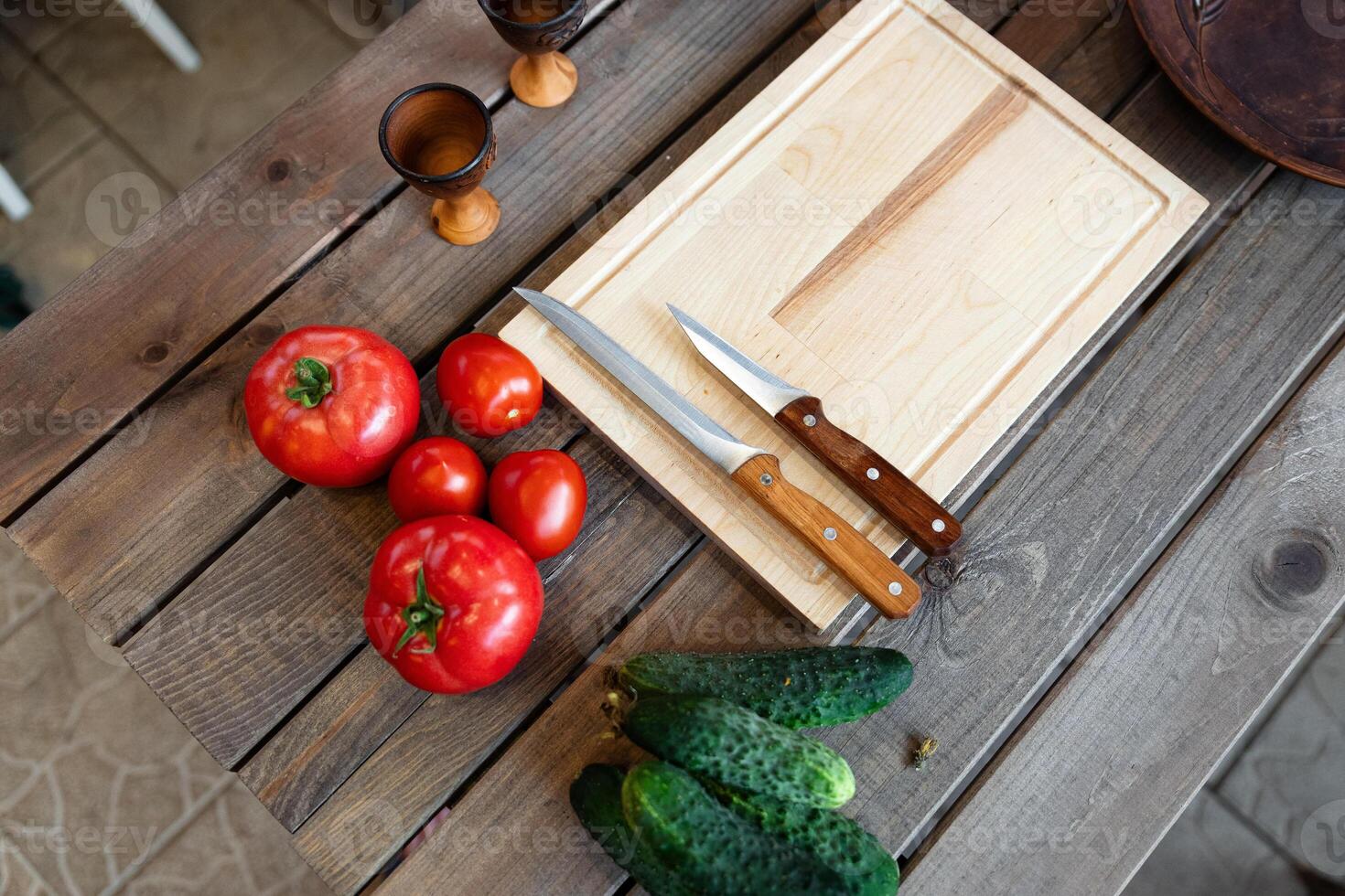 Wooden table with some fresh vegetables and kitchen items. There are several tomatoes and cucumbers placed next to a wooden cutting board. photo