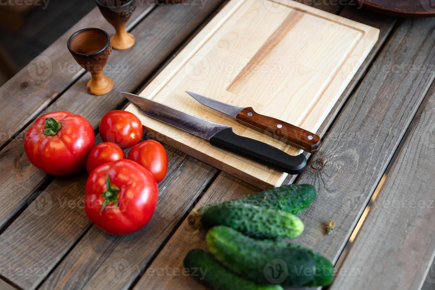 Wooden table with some fresh vegetables and kitchen items. There are several tomatoes and cucumbers placed next to a wooden cutting board. photo