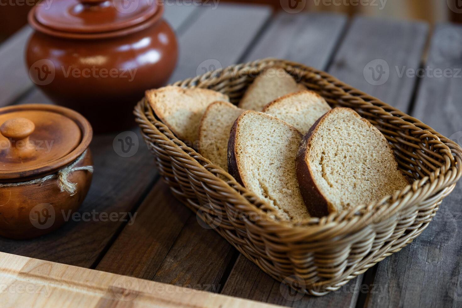 Basket filled with slices of bread. The basket is woven and sits on a wooden table. There are also two ceramic pots with lids nearby, adding a rustic feel to the scene. photo
