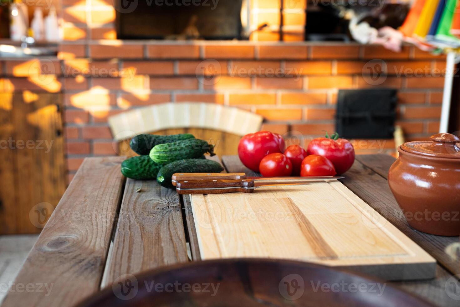 Cutting board on a wooden table, surrounded by fresh vegetables like tomatoes and cucumbers. Rustic kitchen with natural lighting and a brick backdrop. photo