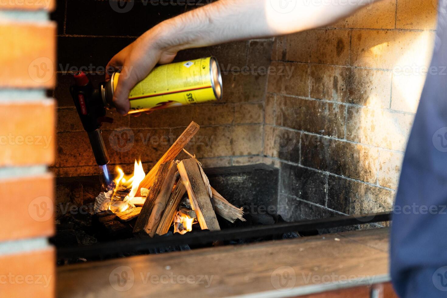 This image shows a fireplace being lit using a butane torch. The flame from the torch is helping ignite pieces of wood arranged in the fireplace. photo