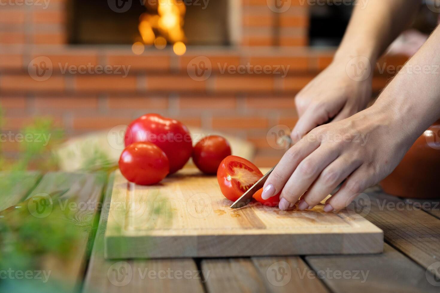 Cutting tomatoes on a wooden cutting board. Cozy kitchen with a brick oven or fireplace in the background, adding warmth. Fresh herbs in the foreground. photo