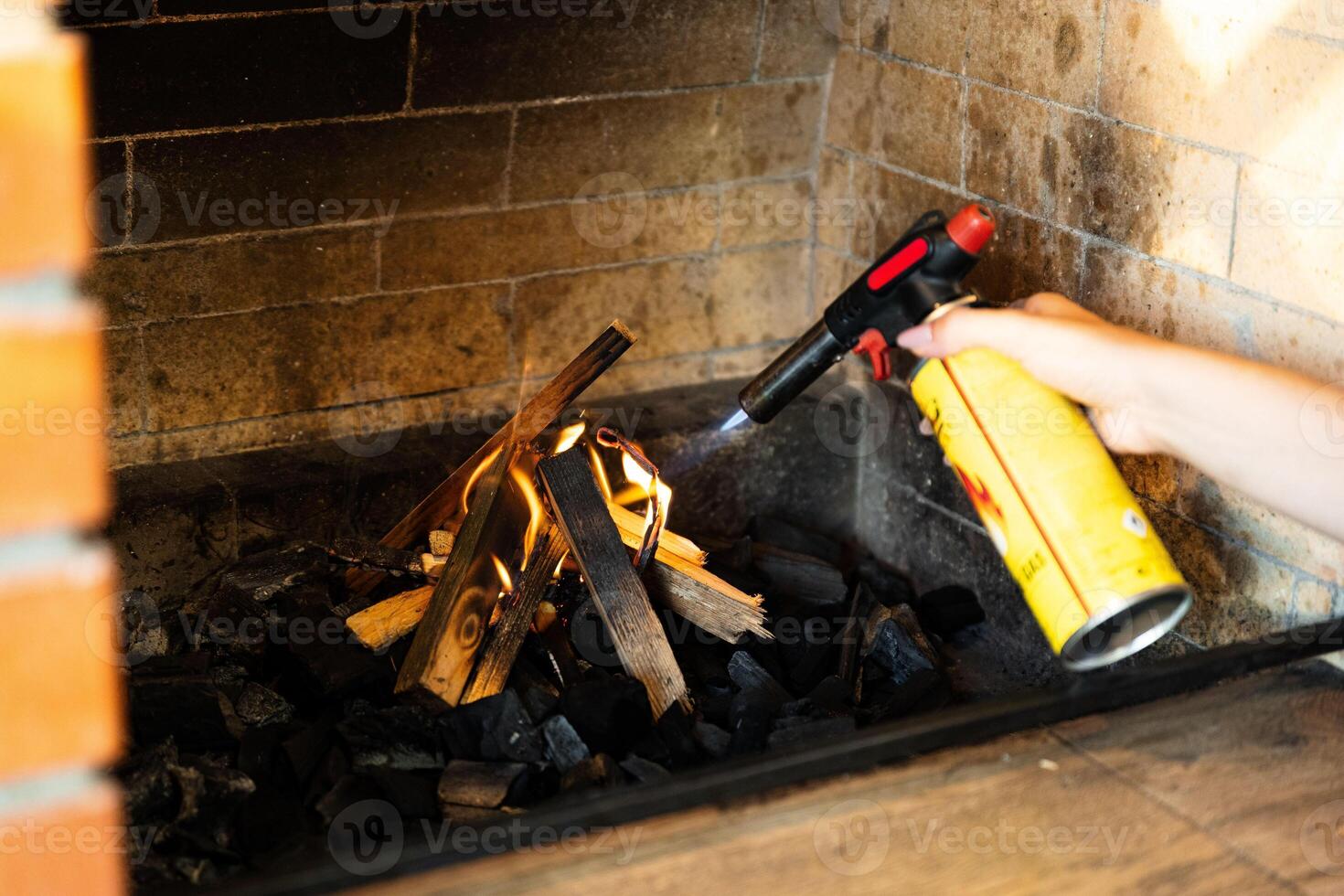 This image shows a fireplace being lit using a butane torch. The flame from the torch is helping ignite pieces of wood arranged in the fireplace. photo