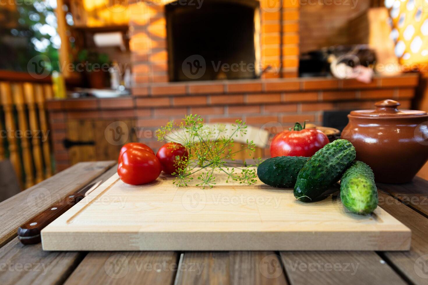 Cutting board on a wooden table, surrounded by fresh vegetables like tomatoes and cucumbers. Rustic kitchen with natural lighting and a brick backdrop. photo