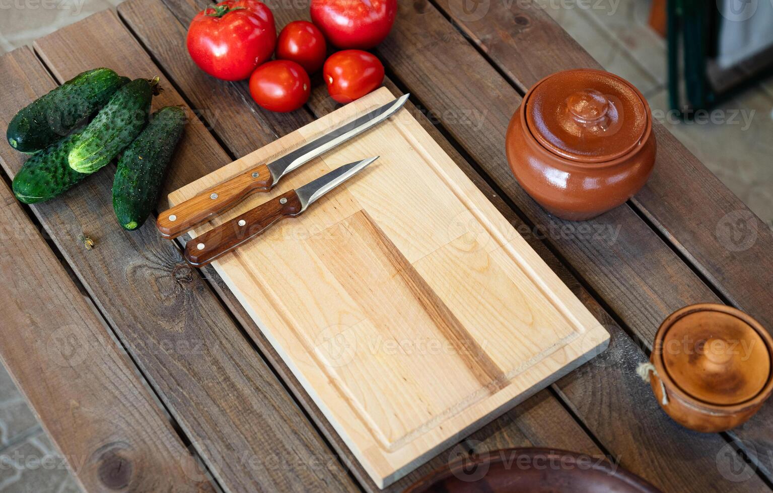 Wooden table with some fresh vegetables and kitchen items. There are several tomatoes and cucumbers placed next to a wooden cutting board. photo