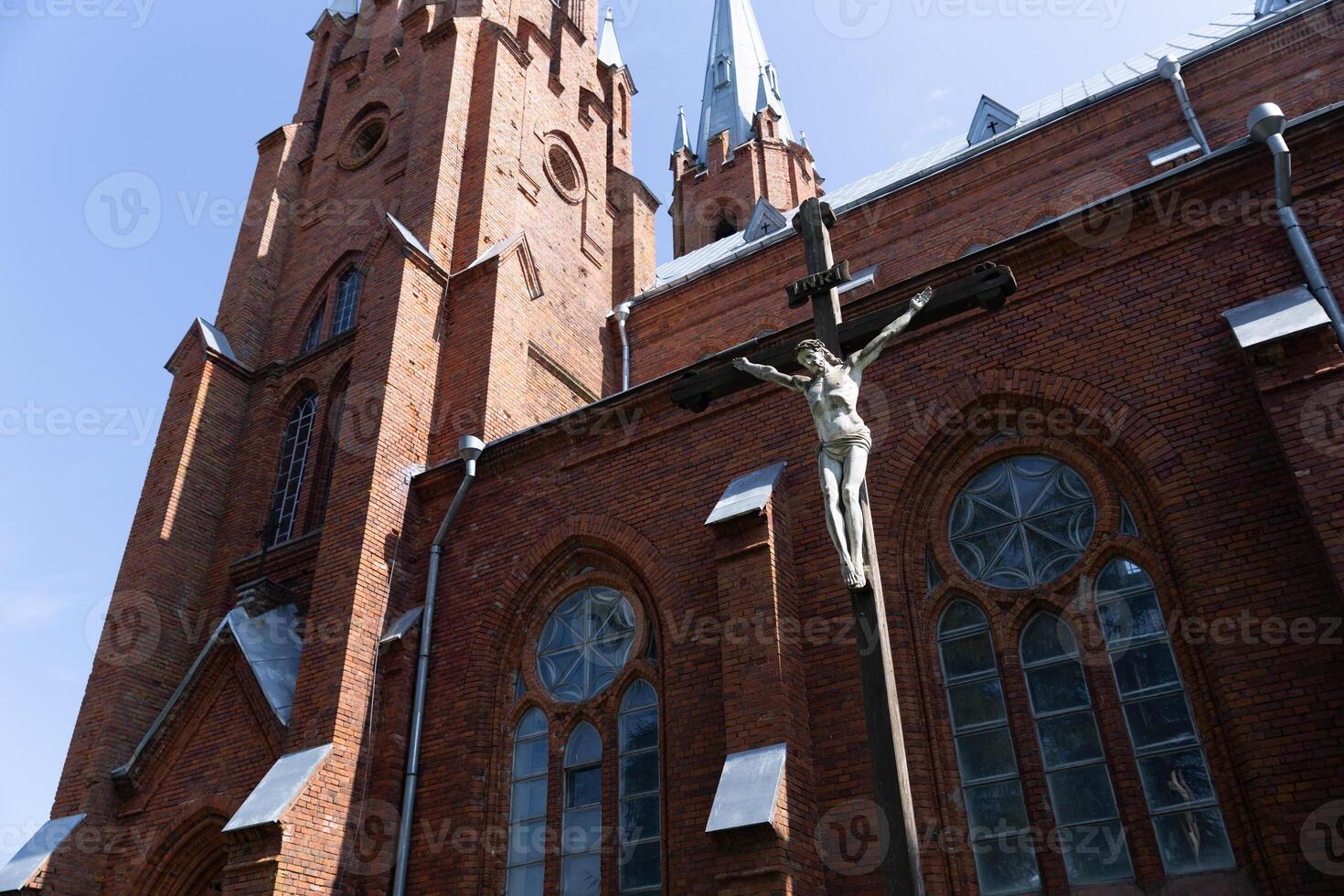 Exterior of a Gothic-style church, with its red brick facade and tall, pointed spires that reach into the sky. Prominently featured is a crucifix, symbolizing the crucifixion of Jesus Christ. photo