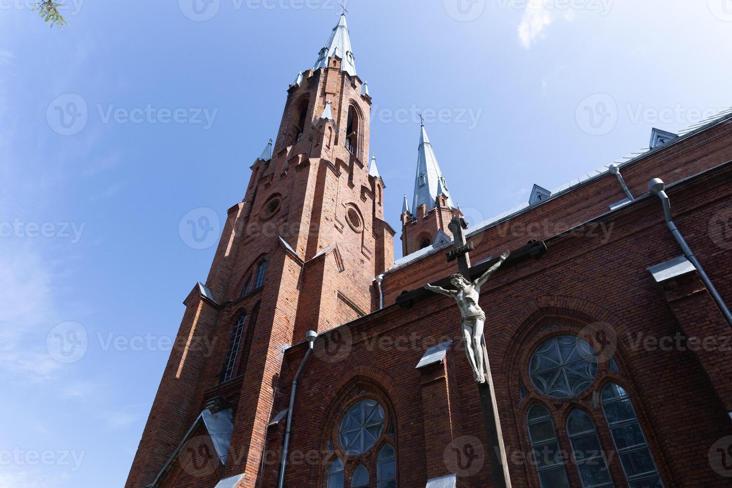 Exterior of a Gothic-style church, with its red brick facade and tall, pointed spires that reach into the sky. Prominently featured is a crucifix, symbolizing the crucifixion of Jesus Christ. photo