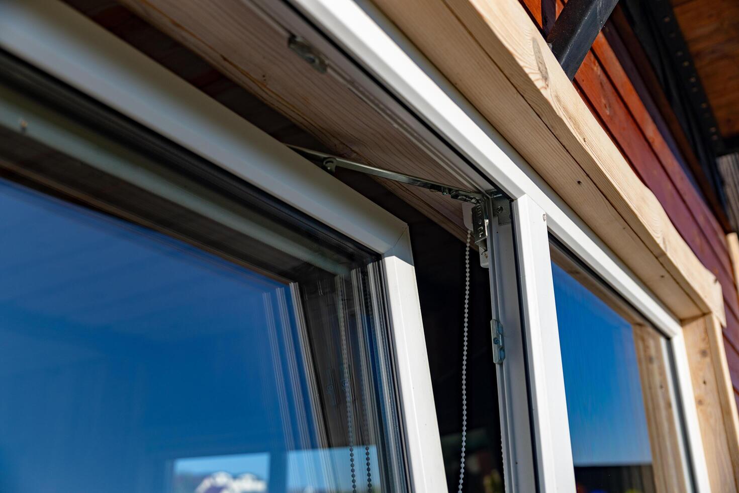 Close-up of a white window frame with a metal hinge and a chain mechanism. The window is open, revealing a view of blue sky. The image captures the details of the window's construction. photo