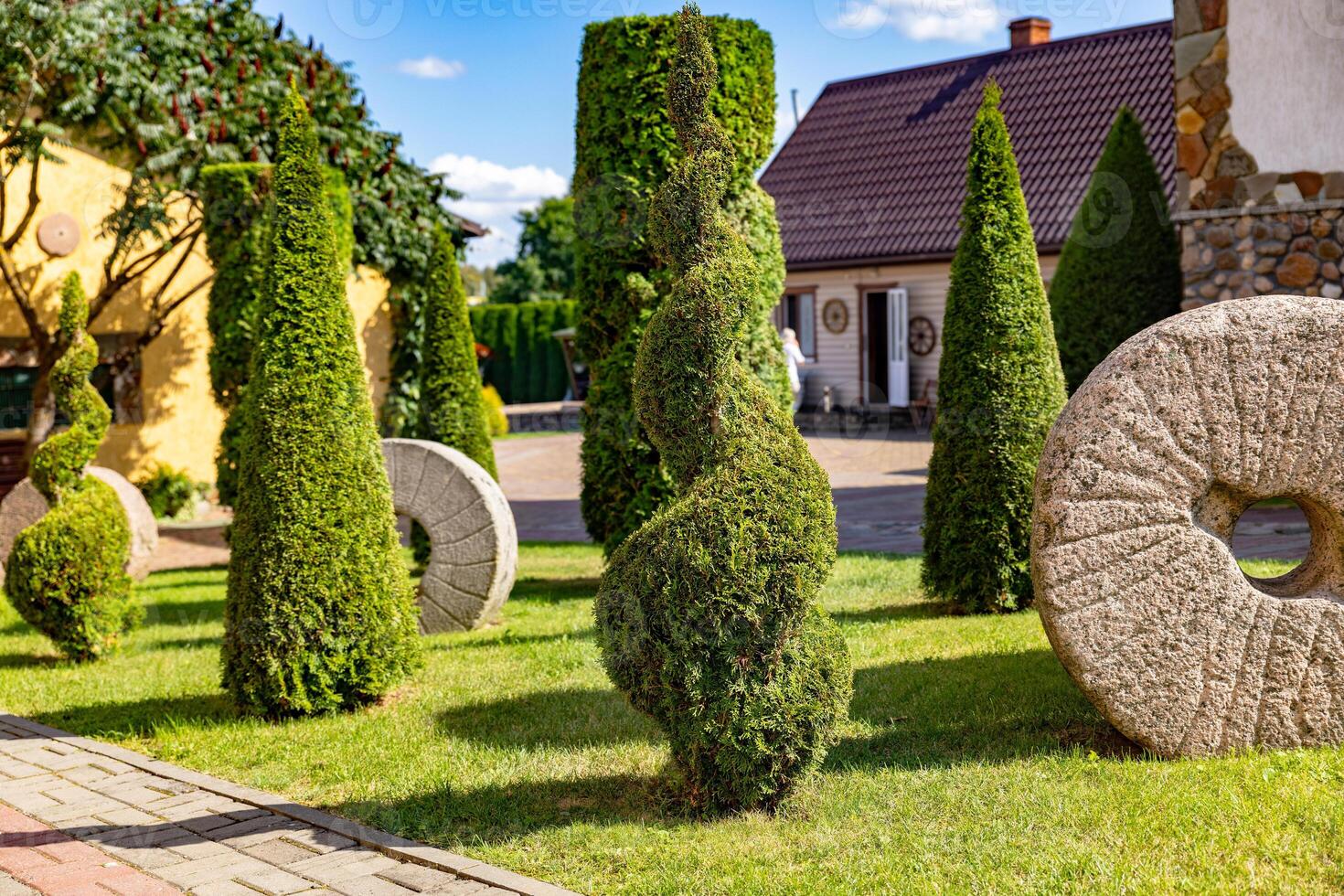 Well-kept garden with a variety of topiary shrubs and a decorative stone mill wheel. With a brick pathway leading into the center of the garden. The trees and shrubs have been carefully shaped. photo
