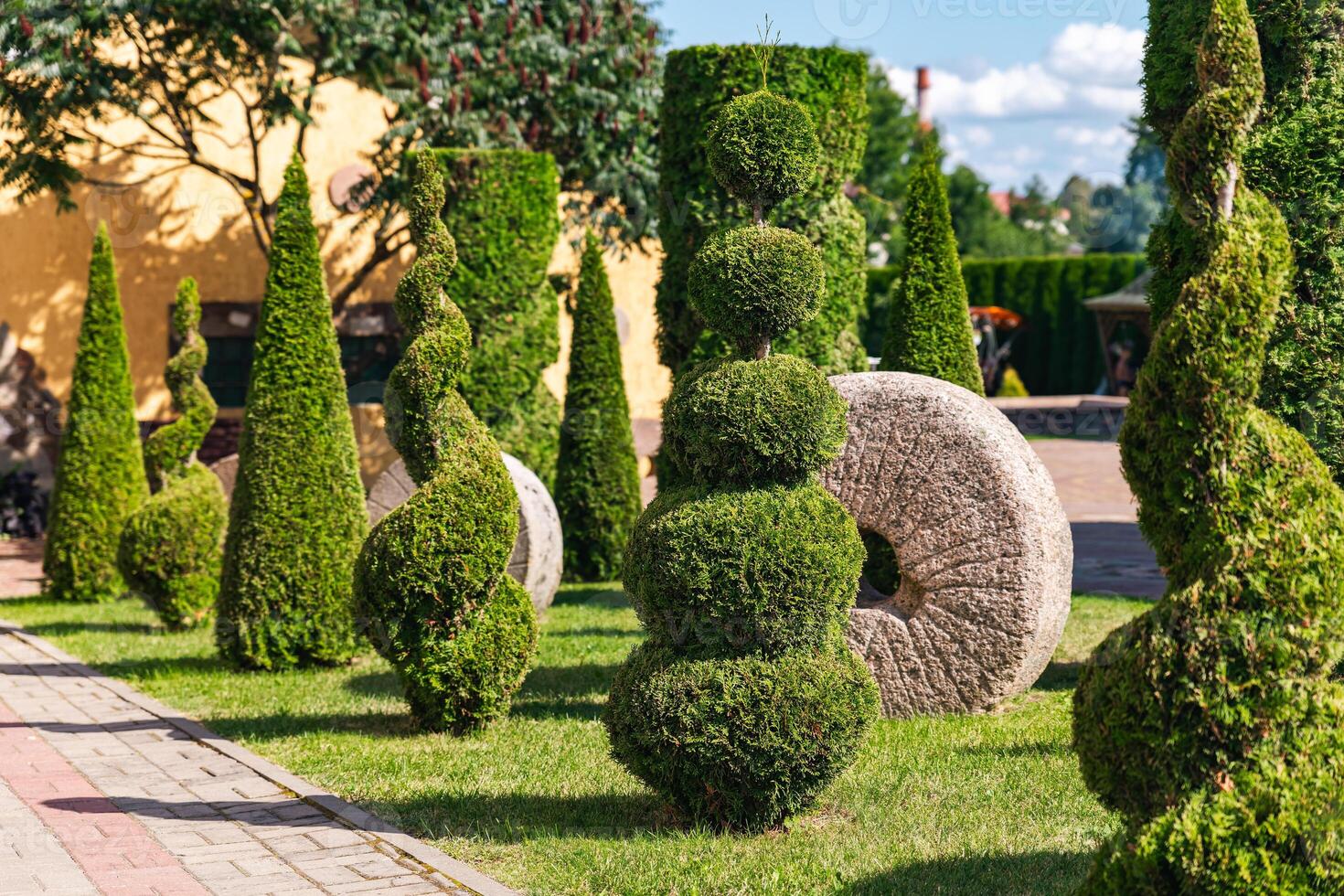 Well-kept garden with a variety of topiary shrubs and a decorative stone mill wheel. With a brick pathway leading into the center of the garden. The trees and shrubs have been carefully shaped. photo