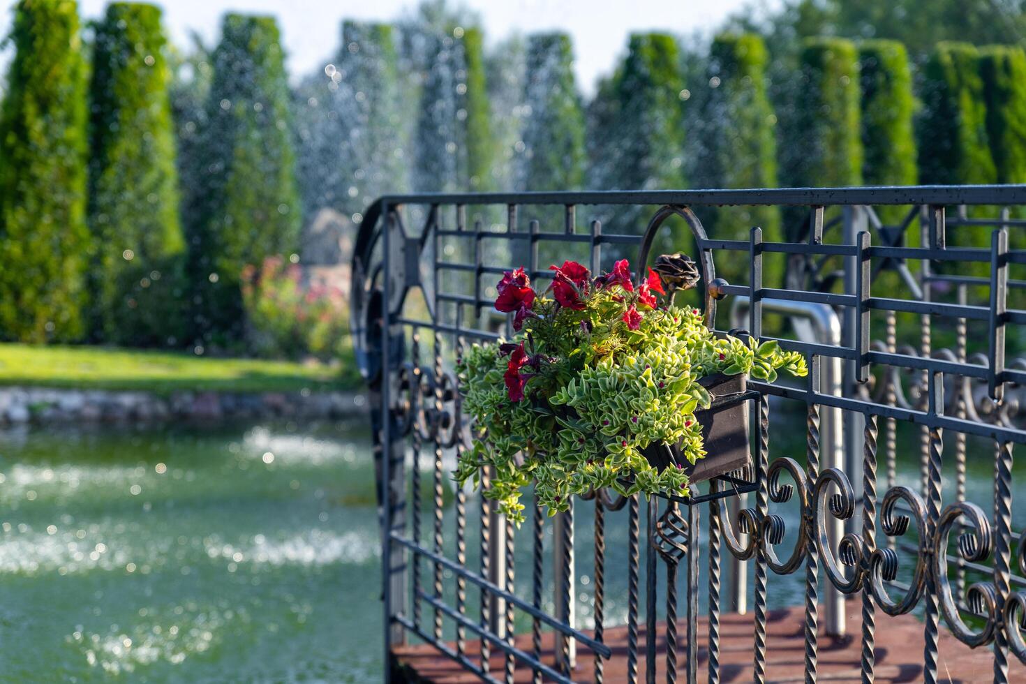 Flowerpot hanging on a decorative iron fence. The background features a blurred view of a pond with a fountain, lush greenery, and a row of tall evergreen trees. Landscape design. photo