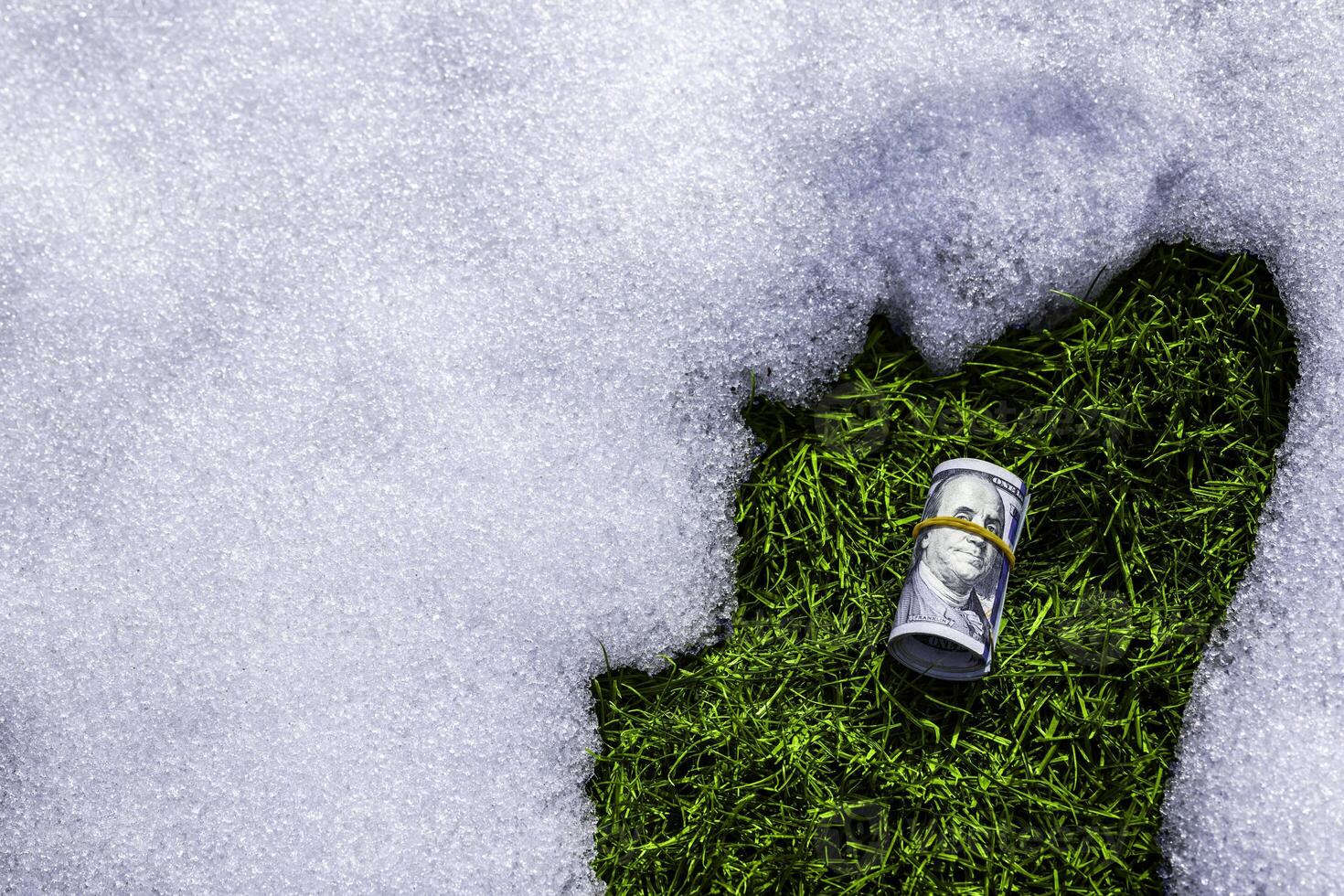 roll of us dollar banknotes on thawed green grass patch in the snow photo