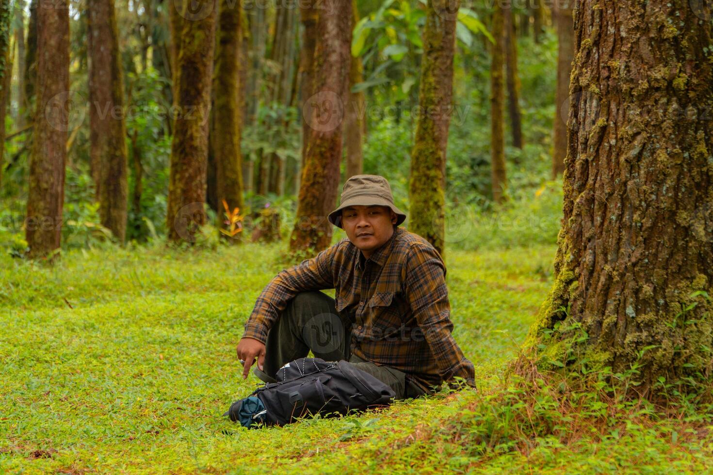 Asian man wearing flannel shirt, bucket hat is sitting and resting while enjoying the tranquility of nature. An adventurer is sitting daydreaming on green grass in a lush pine forest area photo