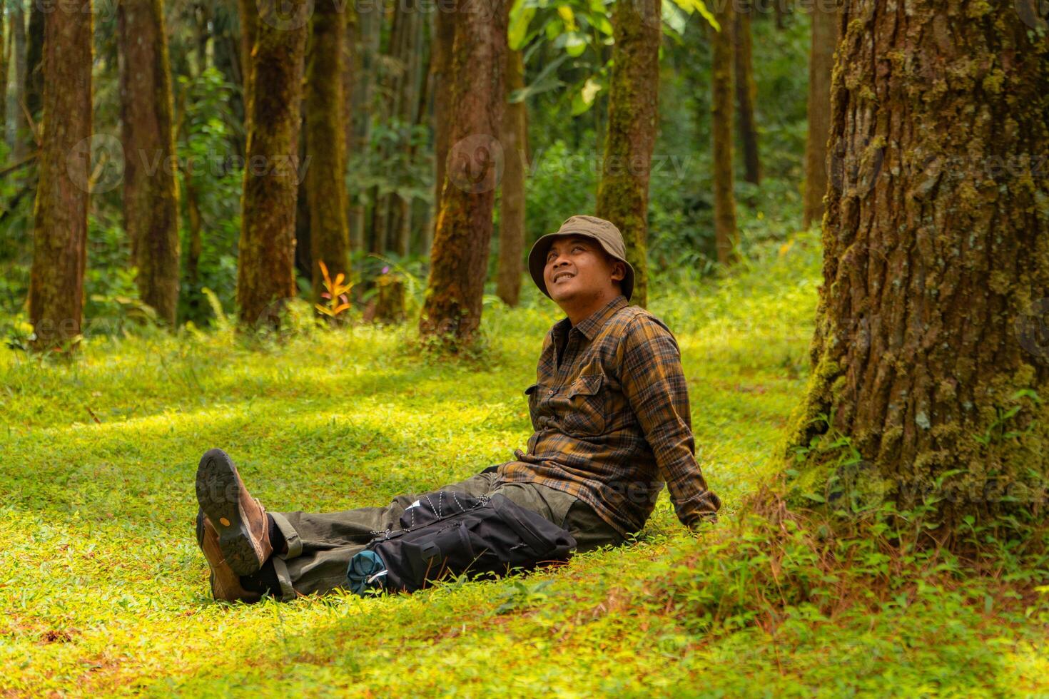 Asian man wearing flannel shirt, bucket hat is sitting and resting while enjoying the tranquility of nature. An adventurer is sitting daydreaming on green grass in a lush pine forest area photo