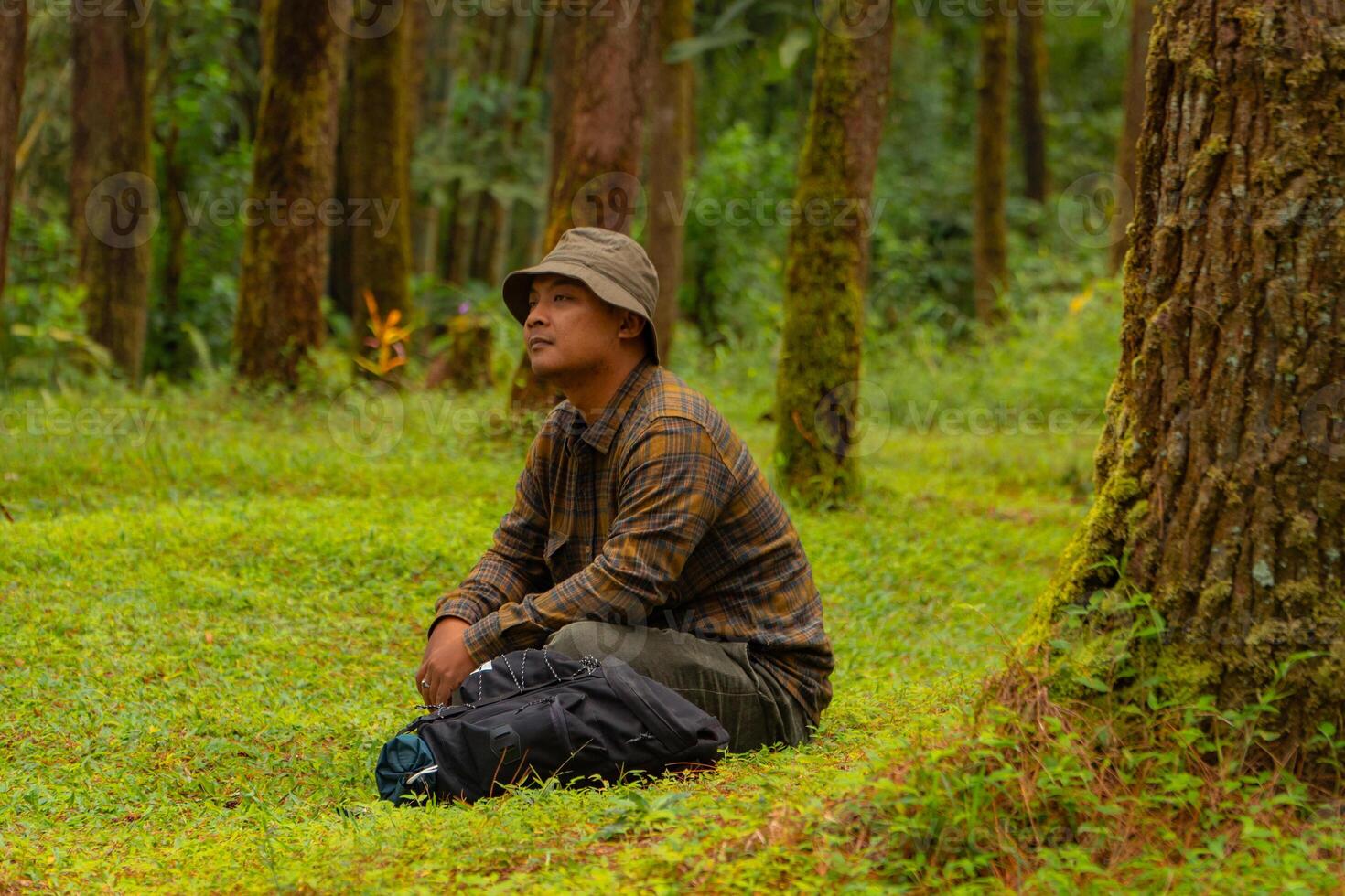 Asian man wearing flannel shirt, bucket hat is sitting and resting while enjoying the tranquility of nature. An adventurer is sitting daydreaming on green grass in a lush pine forest area photo