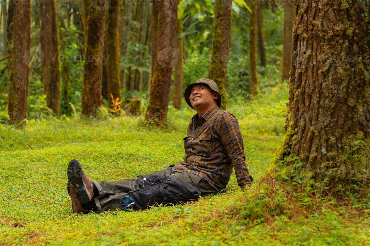 Asian man wearing flannel shirt, bucket hat is sitting and resting while enjoying the tranquility of nature. An adventurer is sitting daydreaming on green grass in a lush pine forest area photo