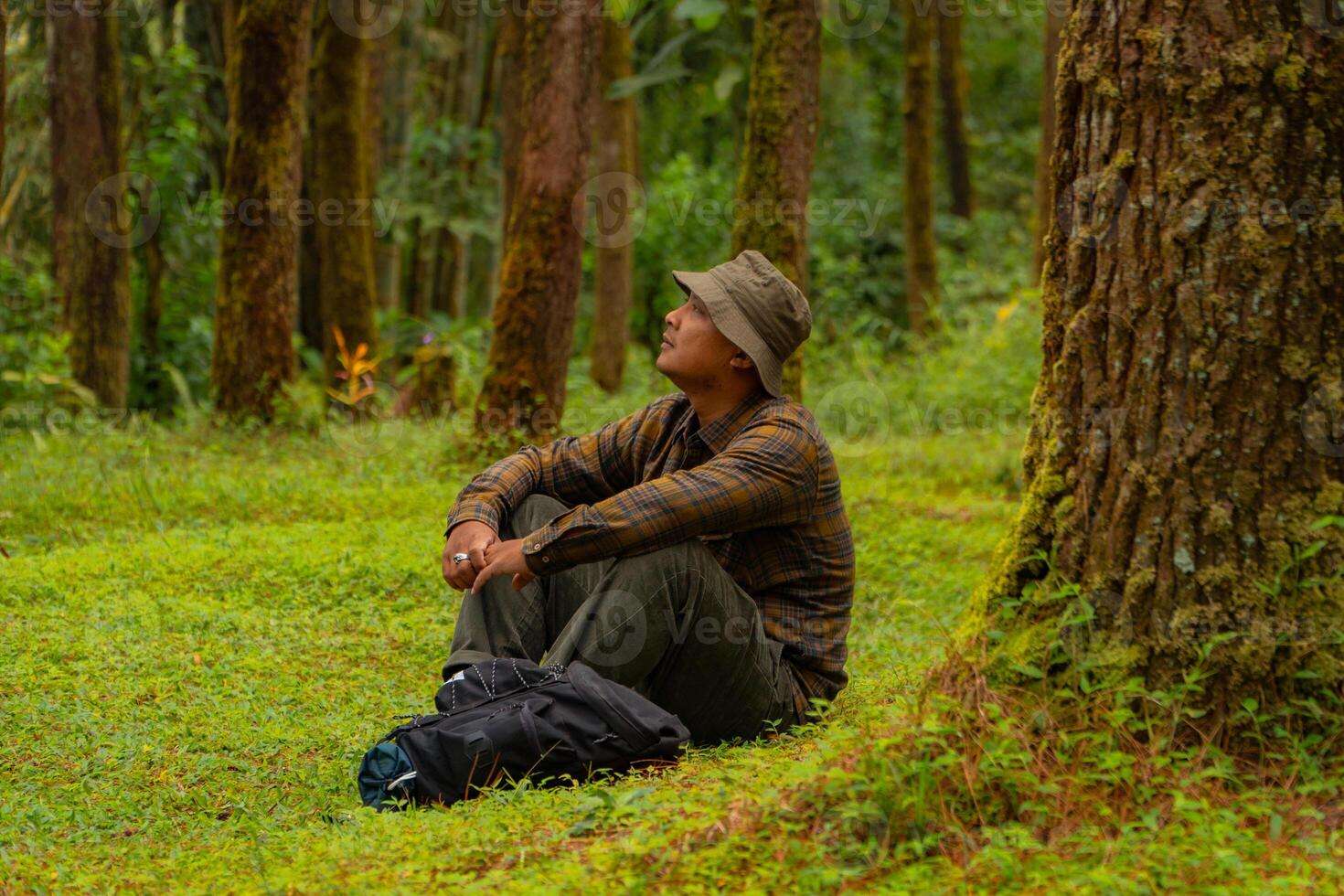 Asian man wearing flannel shirt, bucket hat is sitting and resting while enjoying the tranquility of nature. An adventurer is sitting daydreaming on green grass in a lush pine forest area photo