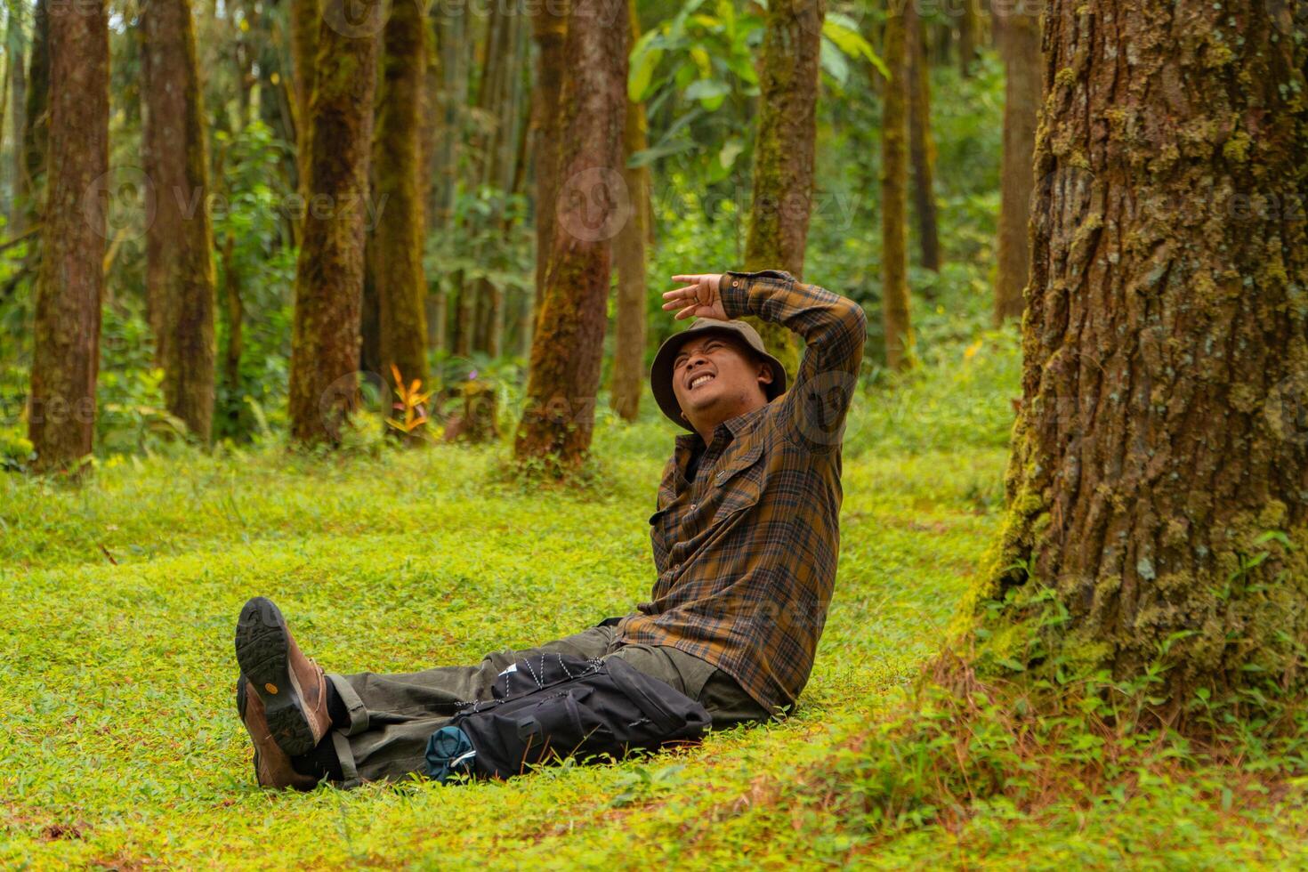 Asian man wearing flannel shirt, bucket hat is sitting and resting while enjoying the tranquility of nature. An adventurer is sitting daydreaming on green grass in a lush pine forest area photo