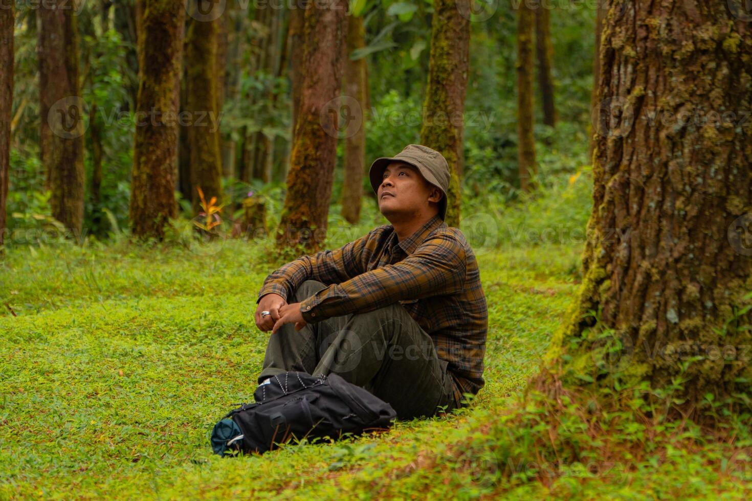 Asian man wearing flannel shirt, bucket hat is sitting and resting while enjoying the tranquility of nature. An adventurer is sitting daydreaming on green grass in a lush pine forest area photo