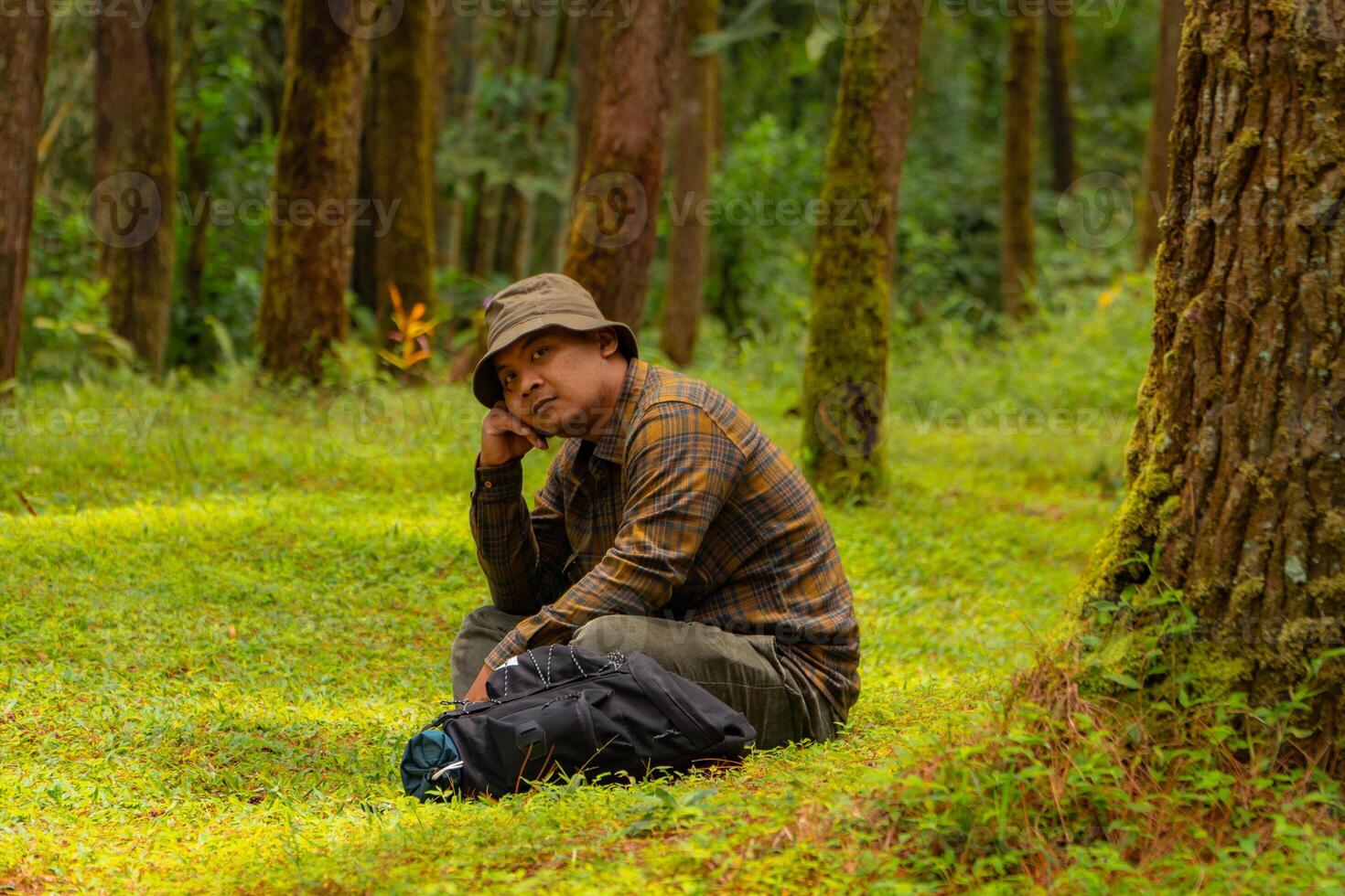 Asian man wearing flannel shirt, bucket hat is sitting and resting while enjoying the tranquility of nature. An adventurer is sitting daydreaming on green grass in a lush pine forest area photo