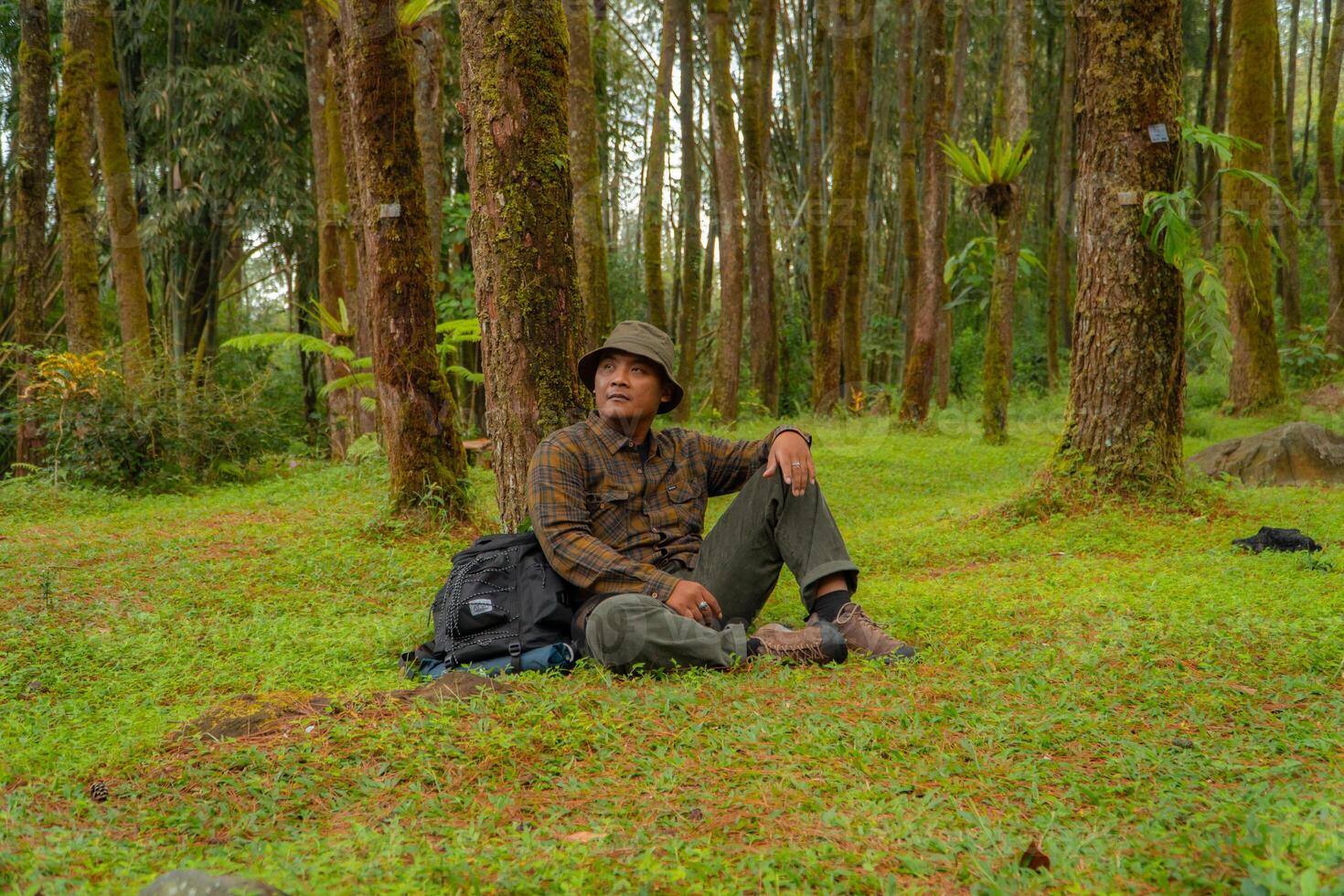 Asian man wearing flannel shirt, bucket hat is sitting and resting while enjoying the tranquility of nature. An adventurer is sitting daydreaming on green grass in a lush pine forest area photo