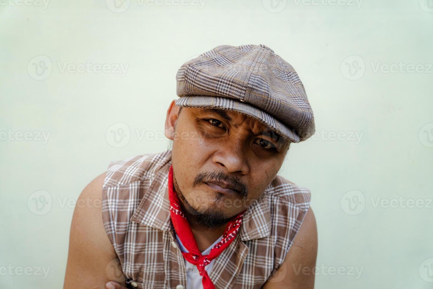 Half body portrait of adult Southeast Asian man in sleeveless shirt, bandana and newsboy hat. Macho male model with beard and mustache looking serious and fierce isolated on beige background. photo