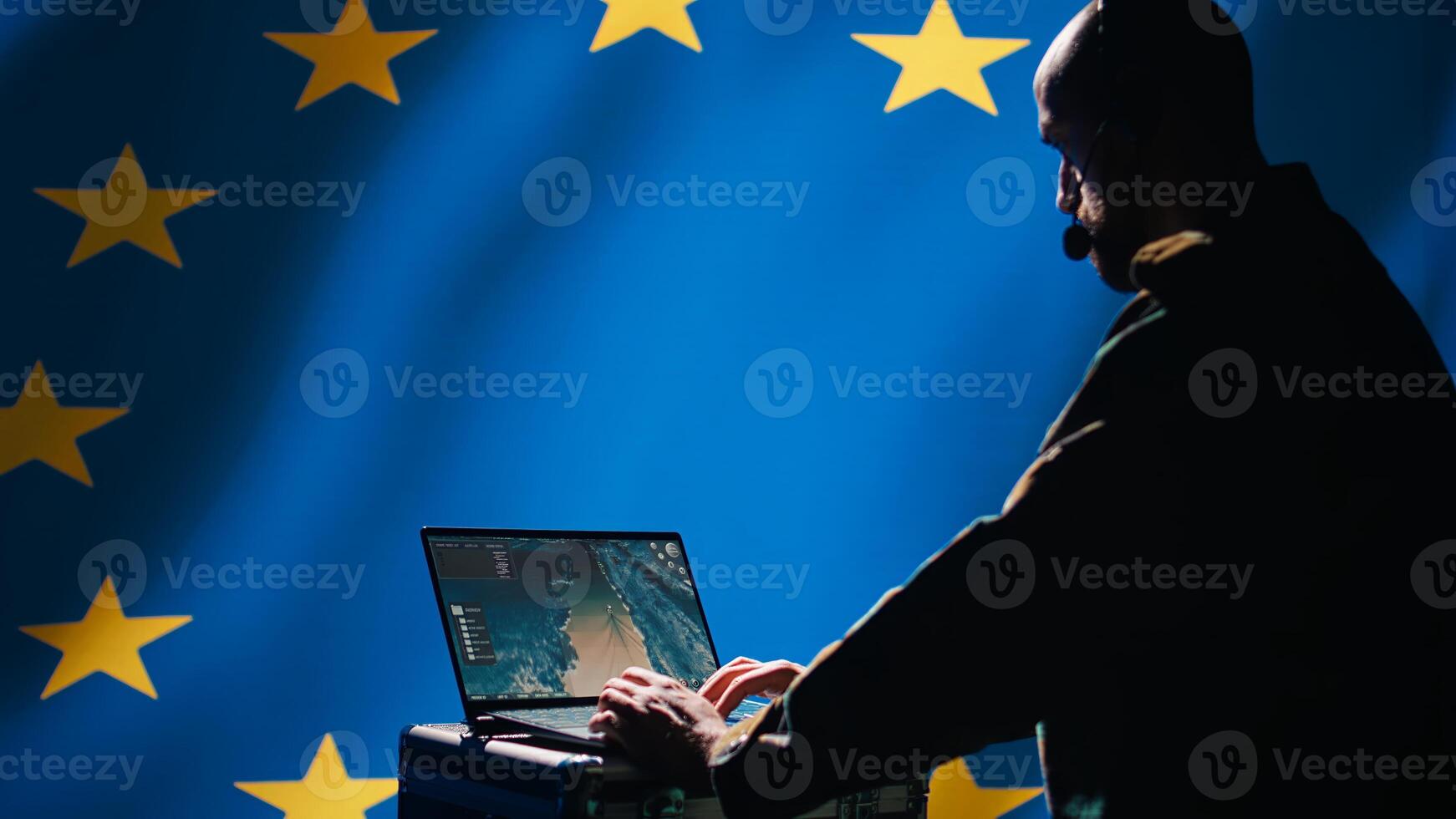 European Union military dispatcher looking at surveillance broadcast, relaying informations for troops using headset. Telecommunications operator over EU flag background, coordinating troops, camera B photo